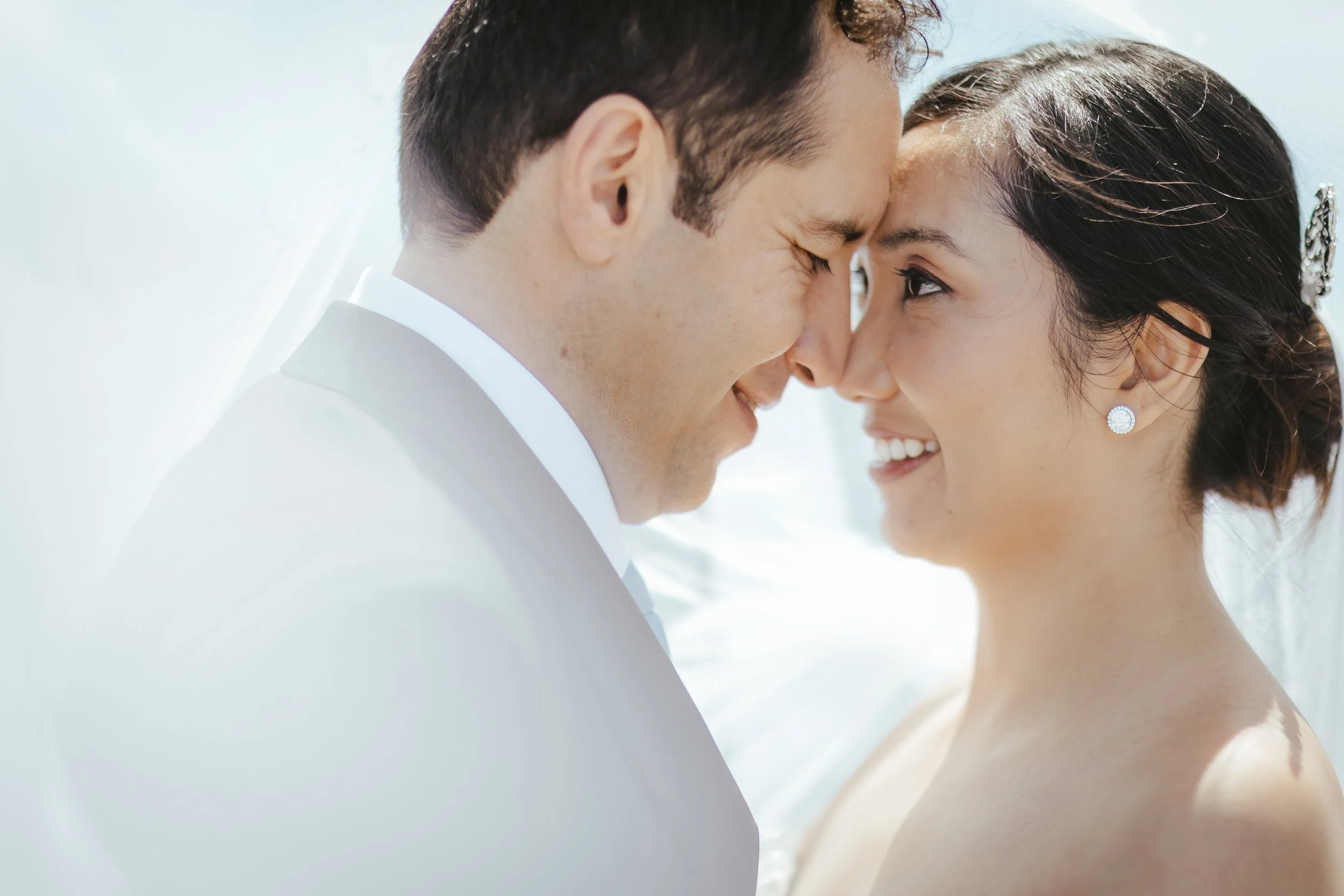 A close-up image of a man and woman with their foreheads touching, smiling and enjoying a moment together. The man has short dark hair, and the woman has dark hair styled with a hair accessory and pearl earrings.