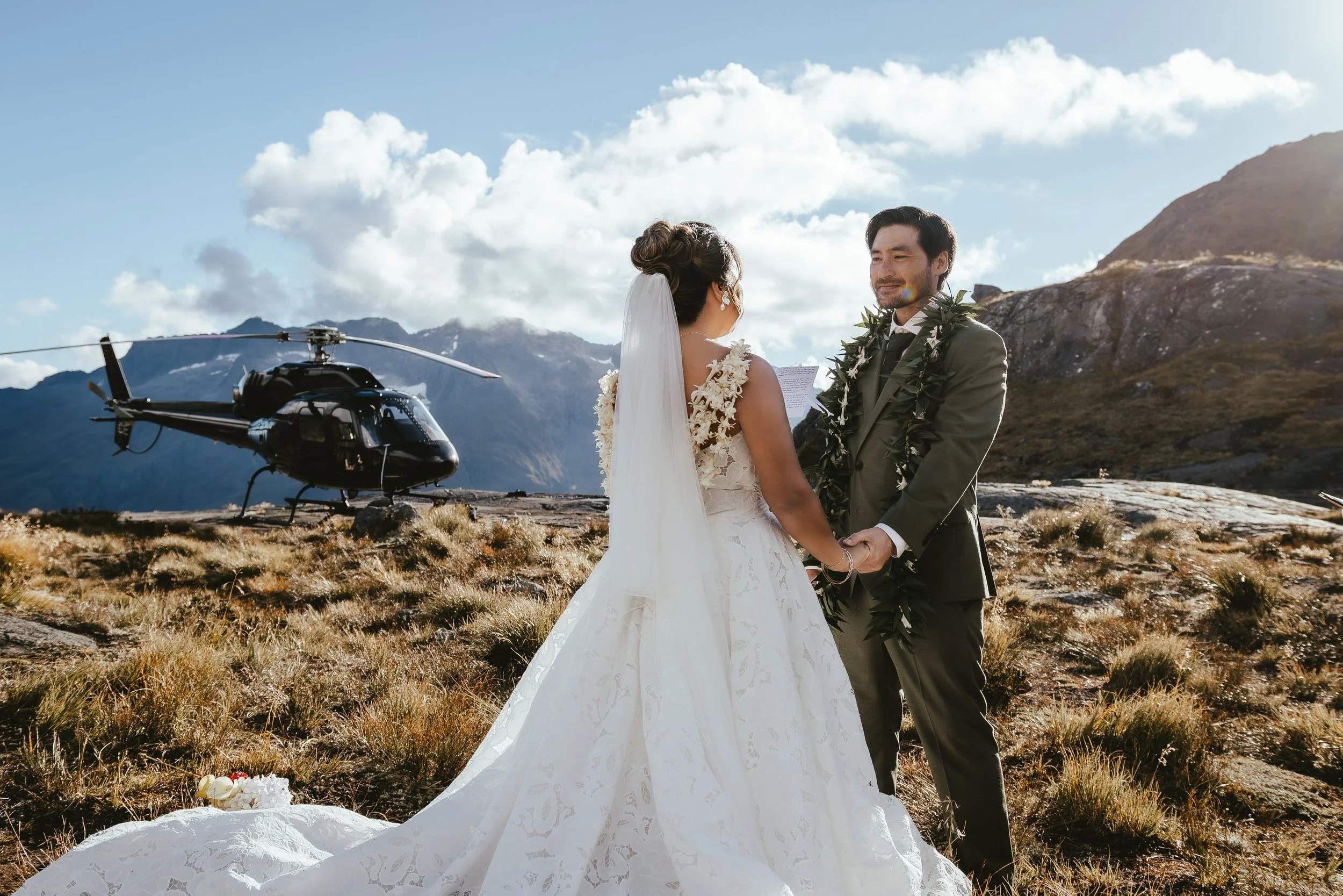 A couple getting married outdoors with mountains in the background, a helicopter nearby, and the bride and groom holding hands.