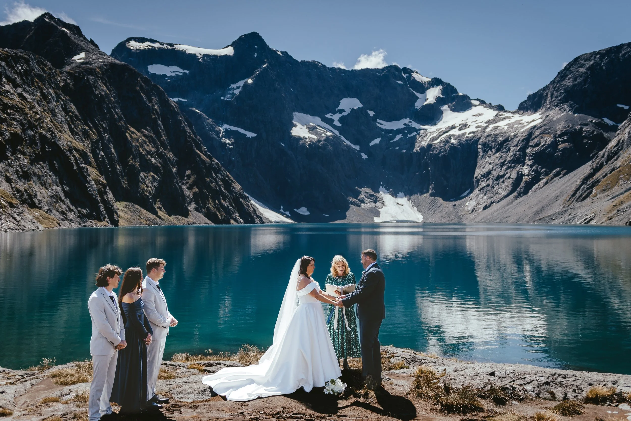 A wedding ceremony taking place outdoors by a lake, with mountains and snow patches in the background. The bride and groom are holding hands, and a officiant is reading from a book. Two of the wedding party members stand to the left.