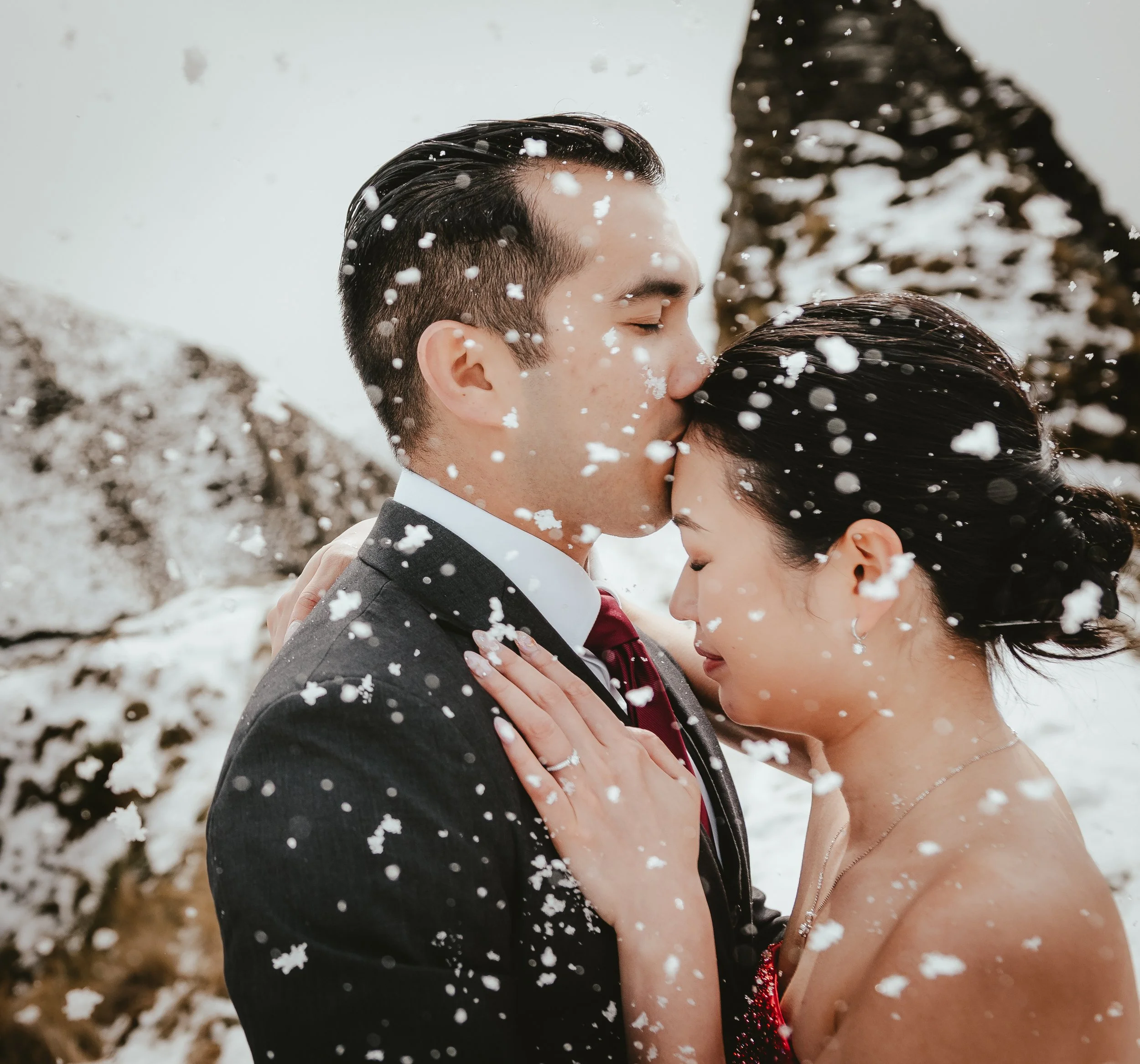 A couple in formal attire, kissing outdoors in a snowy environment. Snowflakes are falling around them.