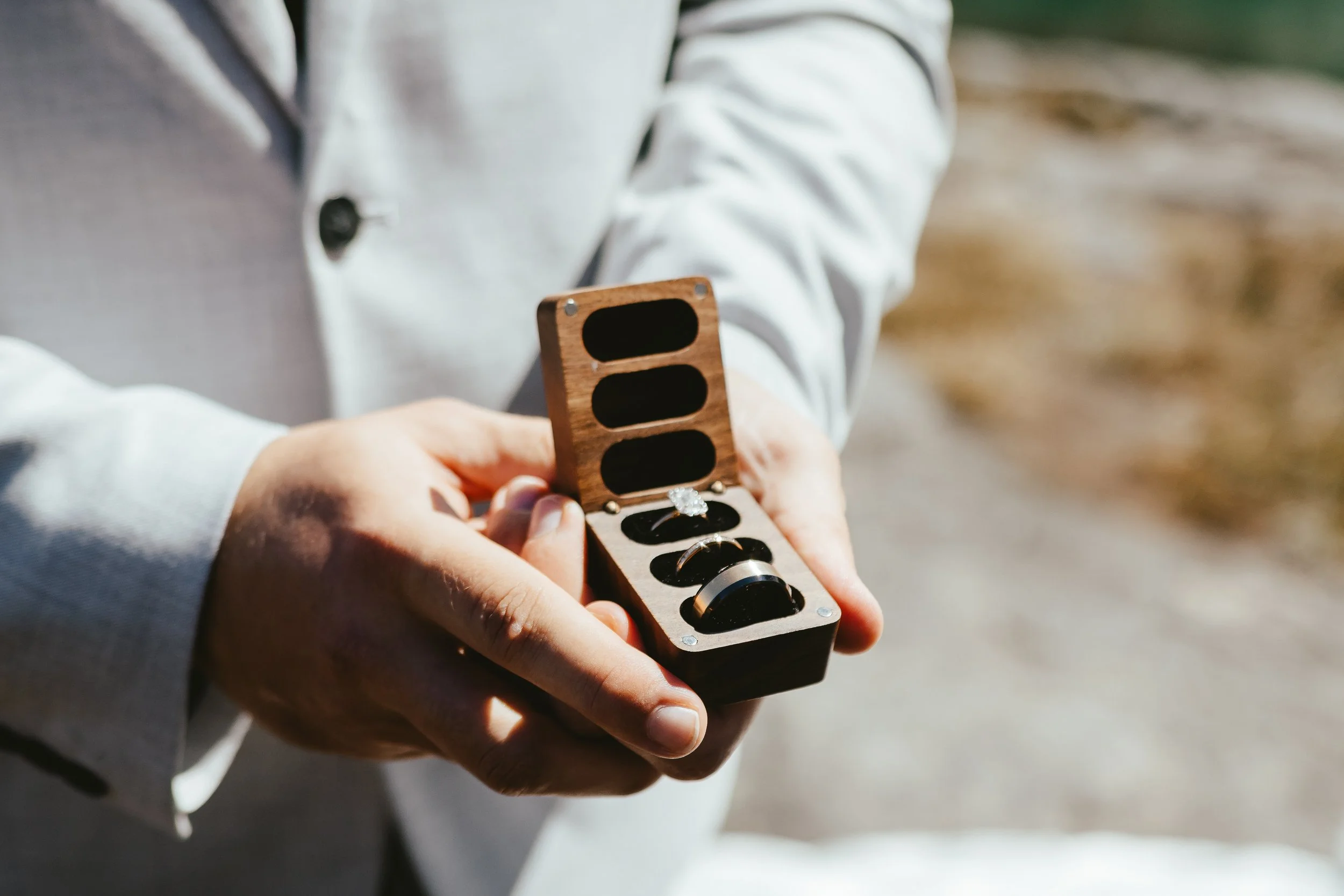 Person holding an open wooden box with rings inside, outdoors.