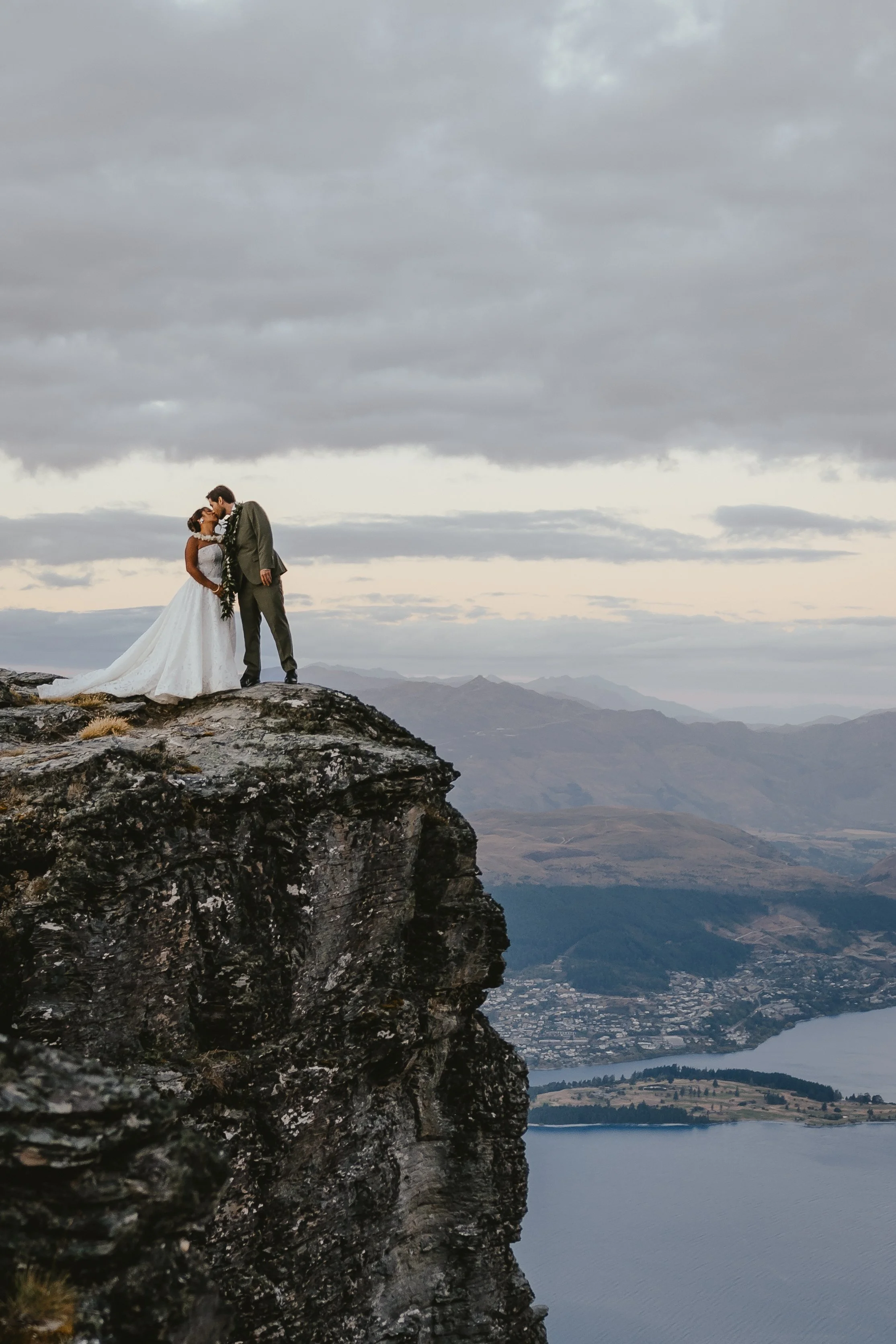 A bride and groom standing close together on a rocky mountain ledge, overlooking a landscape of mountains, water, and a cloudy sky.