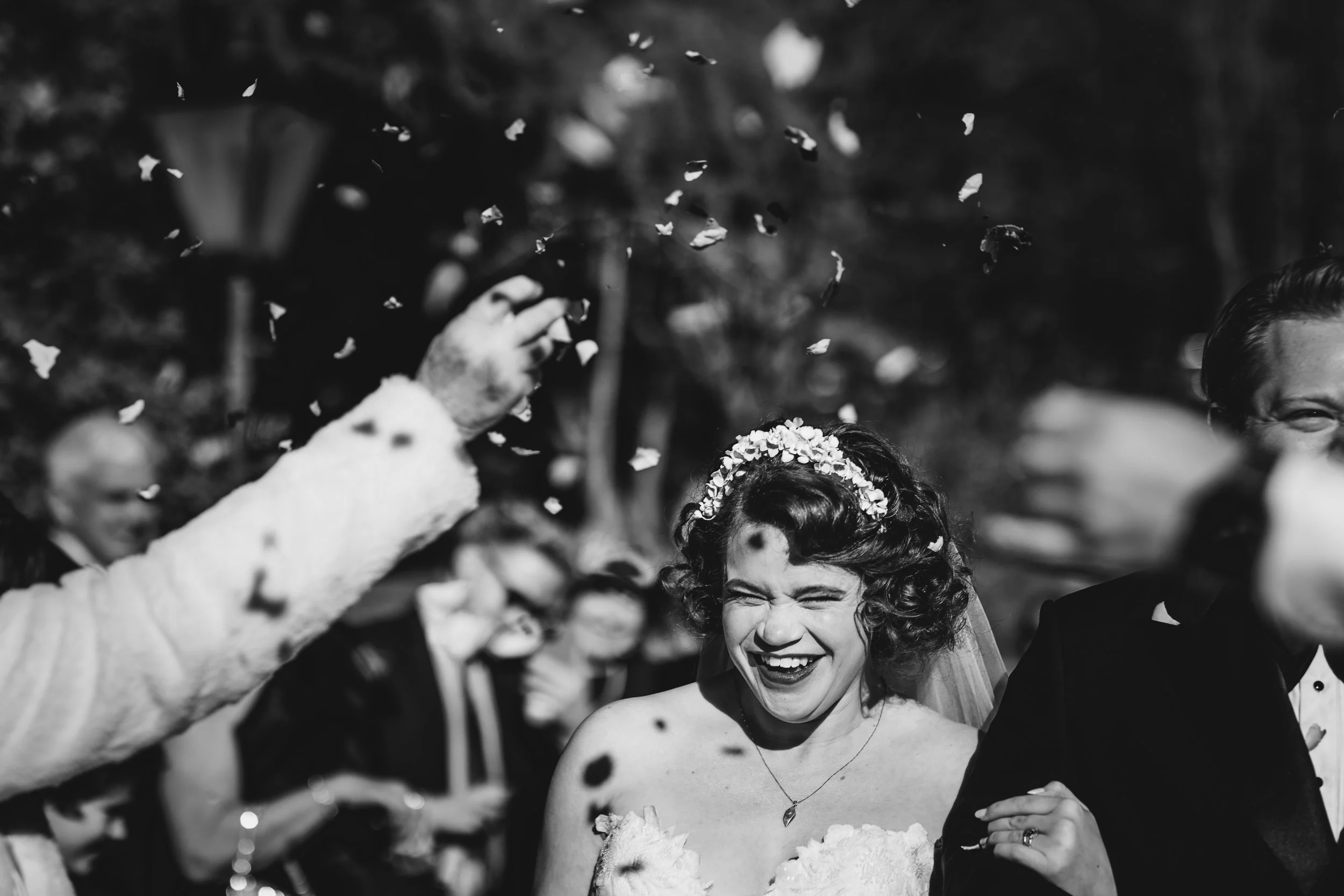 A black and white photo of a bride and groom smiling as they celebrate a wedding. The bride is wearing a floral crown and a dress, and the groom is in a tuxedo. They are surrounded by people throwing confetti in the air, and some guests are visible i
