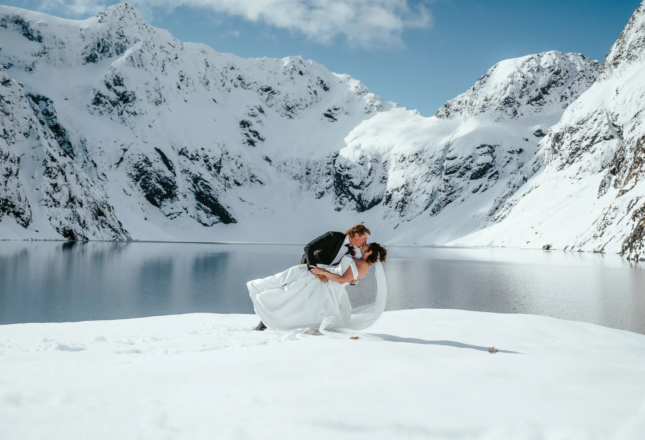 A couple dressed in wedding attire sharing an intimate moment on a snowy landscape, with snow-covered mountains and a clear blue sky in the background.