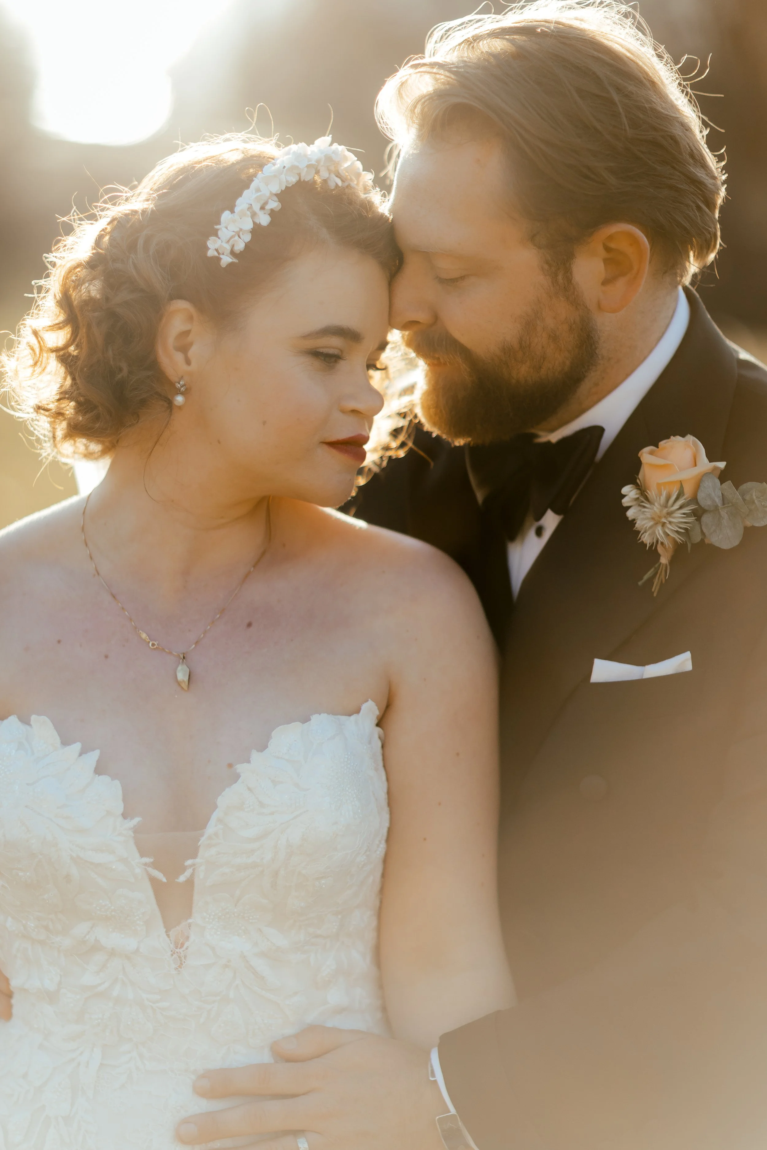 A bride and groom close together, touching foreheads with eyes closed during a wedding ceremony, with warm sunlight in the background.