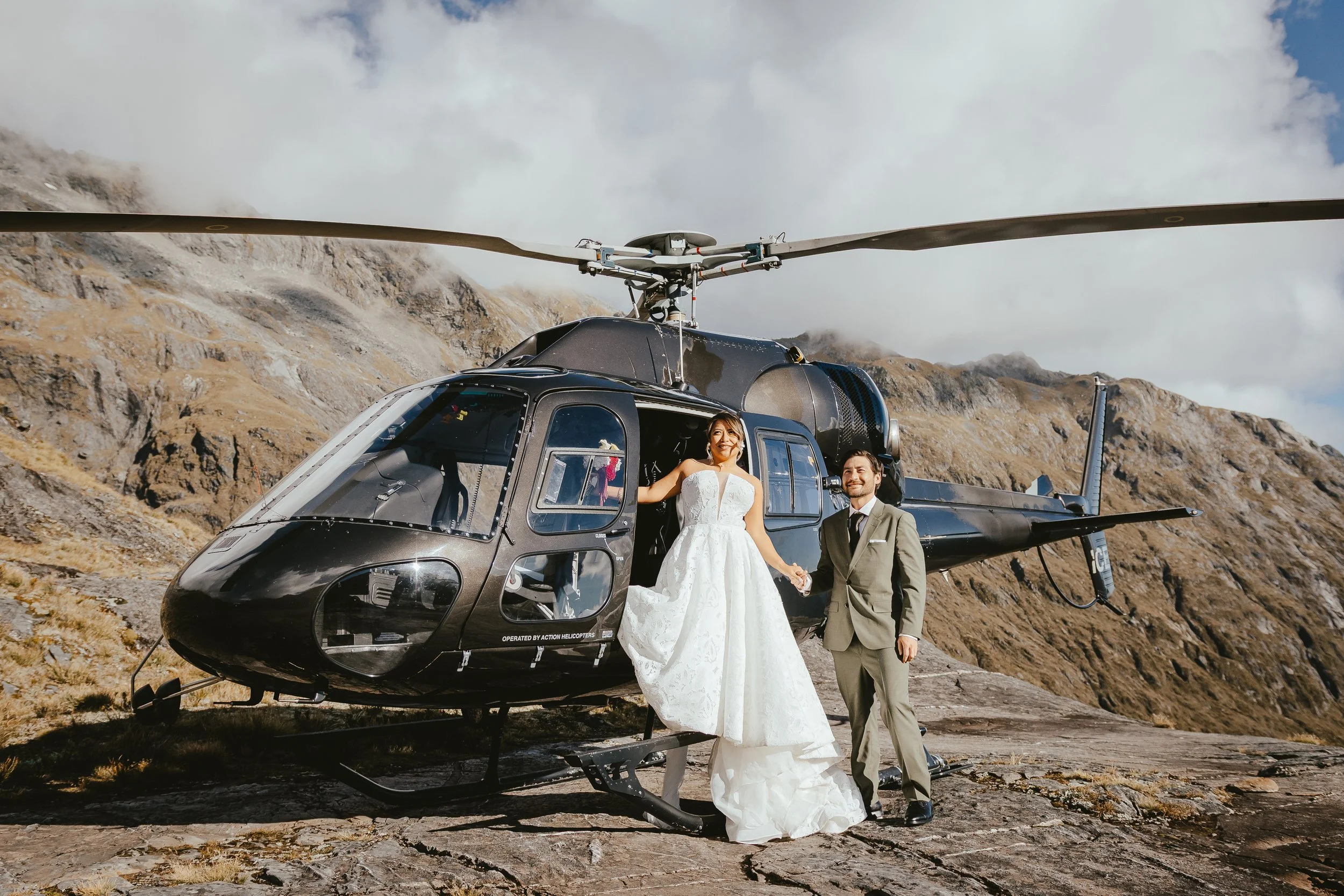 A bride and groom holding hands standing in front of a helicopter in a mountainous landscape with cloudy skies.