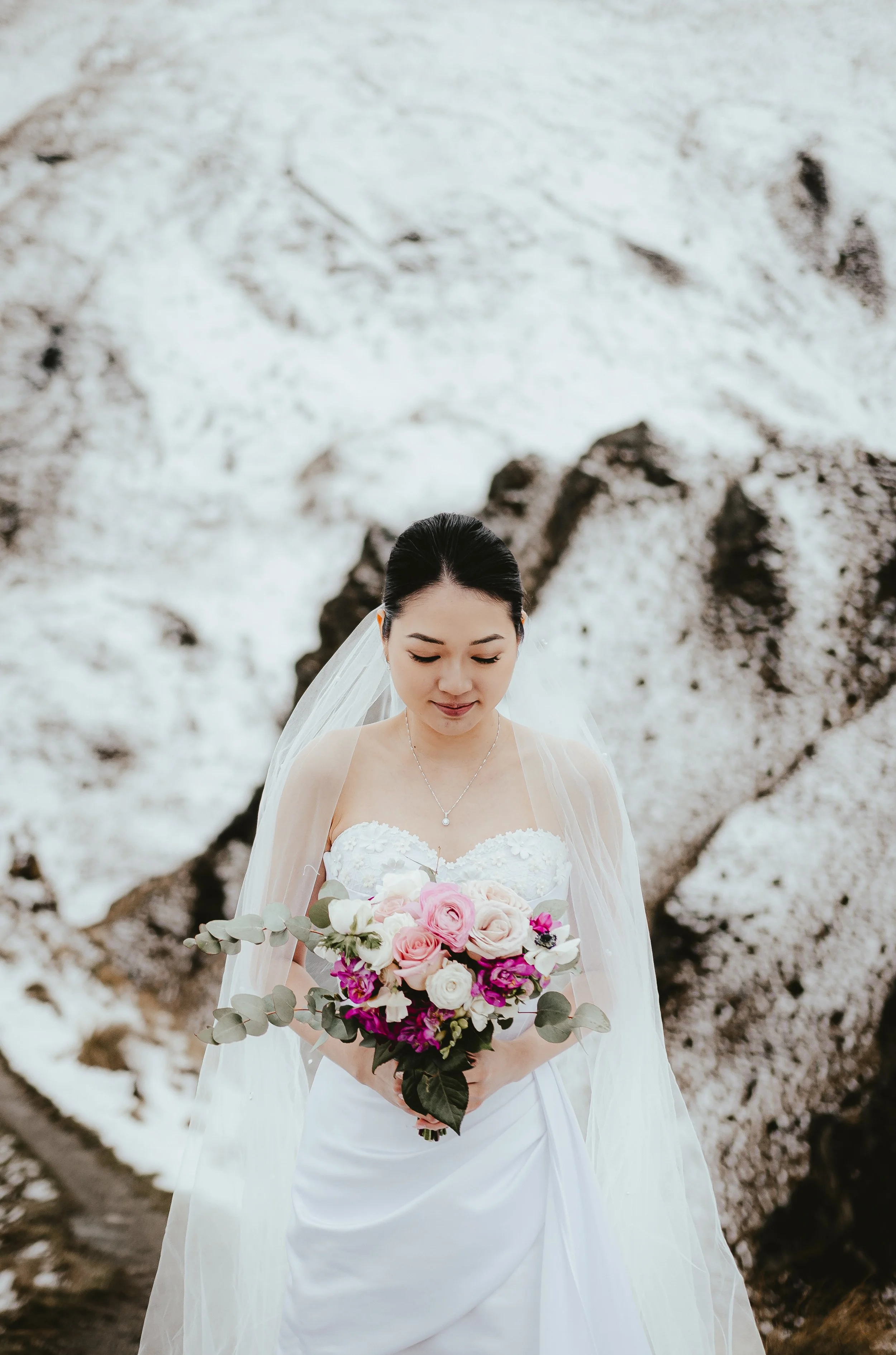 A bride with dark hair in a bun, wearing a white wedding dress with a sheer shoulder overlay, holding a bouquet of pink, white, and purple flowers with greenery, standing outdoors.