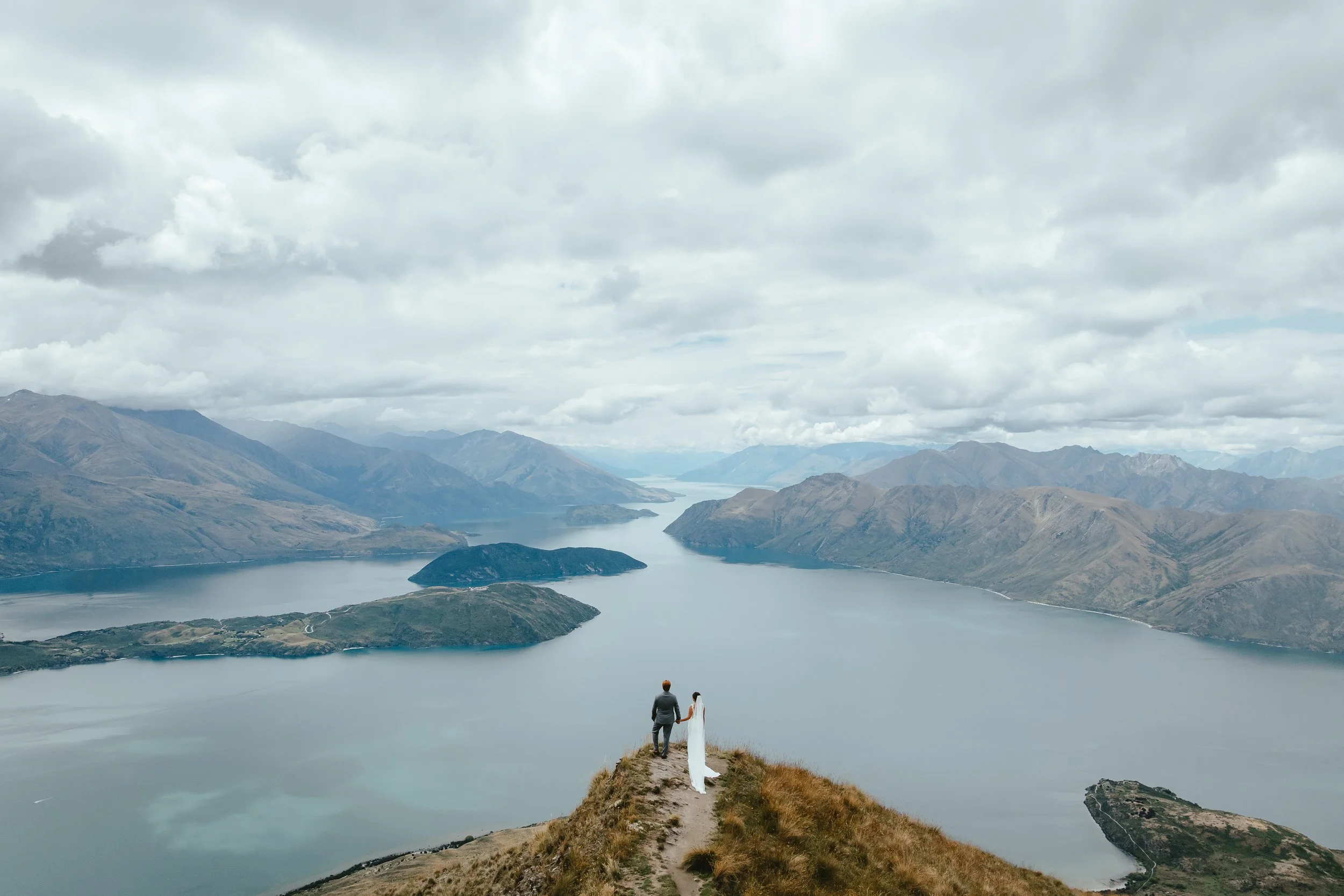A couple walking hand in hand along a narrow path on a hilltop overlooking a large lake or river surrounded by mountains under a cloudy sky.