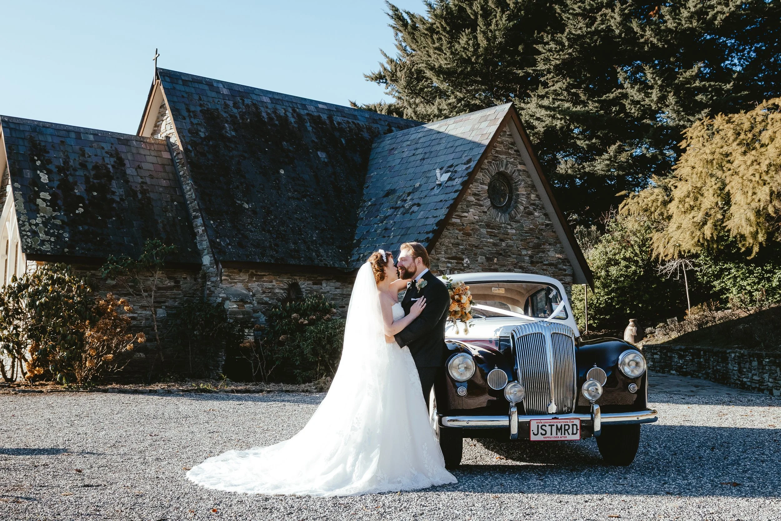A bride and groom in wedding attire sharing a kiss in front of a vintage black car with a custom license plate, standing outside a stone church with a steep roof and a cross on top surrounded by trees.
