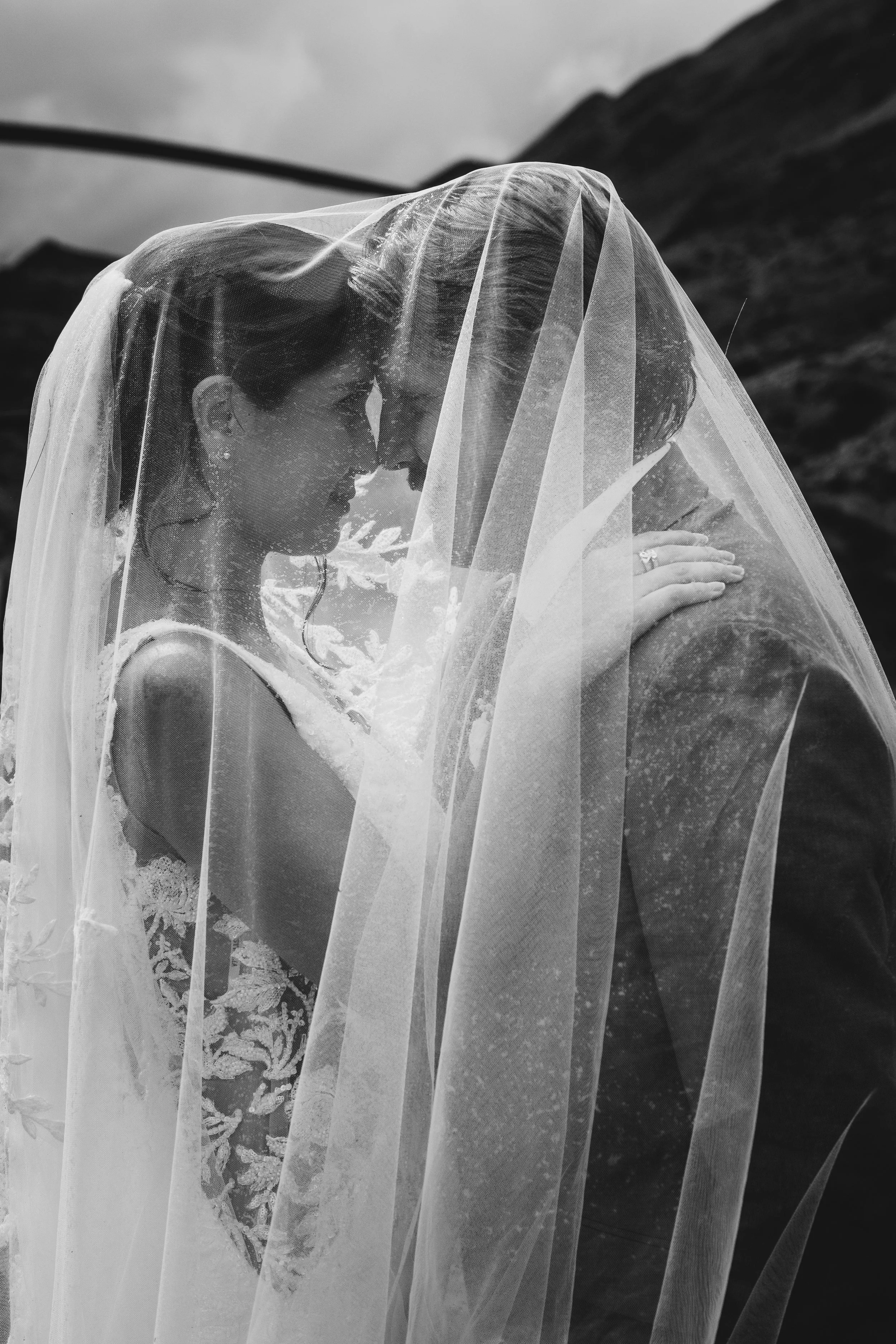 A bride with short hair wearing a lace wedding dress and veil embraces a person outdoors with mountainous terrain in the background.