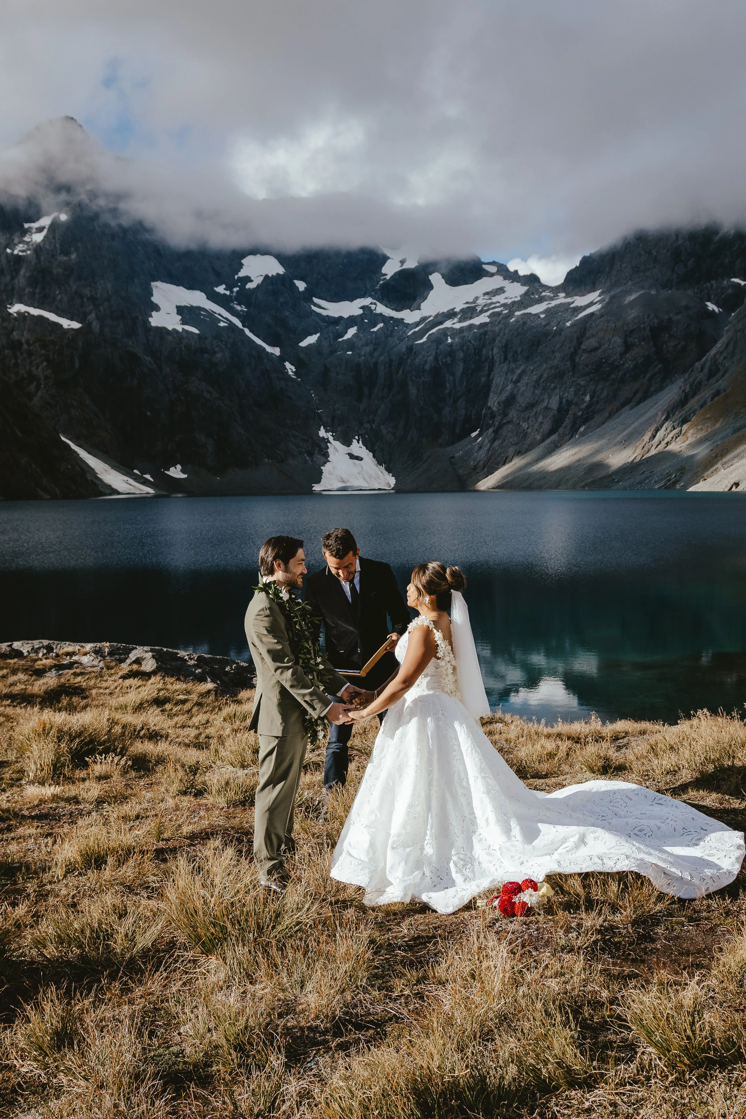 A couple getting married outdoors in a mountainous area by a lake, with an officiant and a person taking photos. The bride is in a white gown with a long train, and the groom is in a suit. The scenery includes snow-capped mountains, a lake, and cloud
