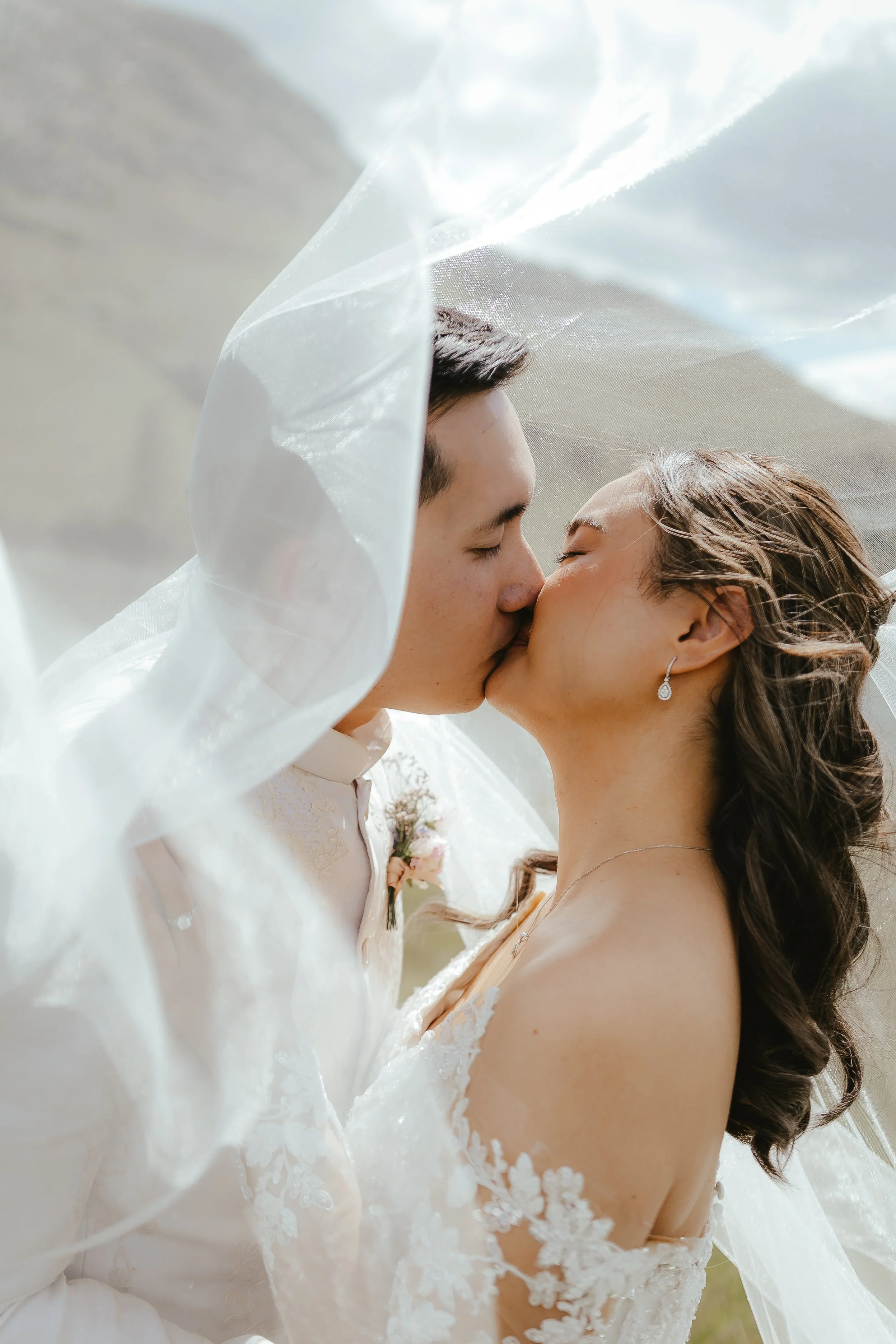 A newlywed couple, a man with closed eyes and a woman wearing earrings, sharing a kiss under a sheer veil with floral lace details, outdoors during daytime.