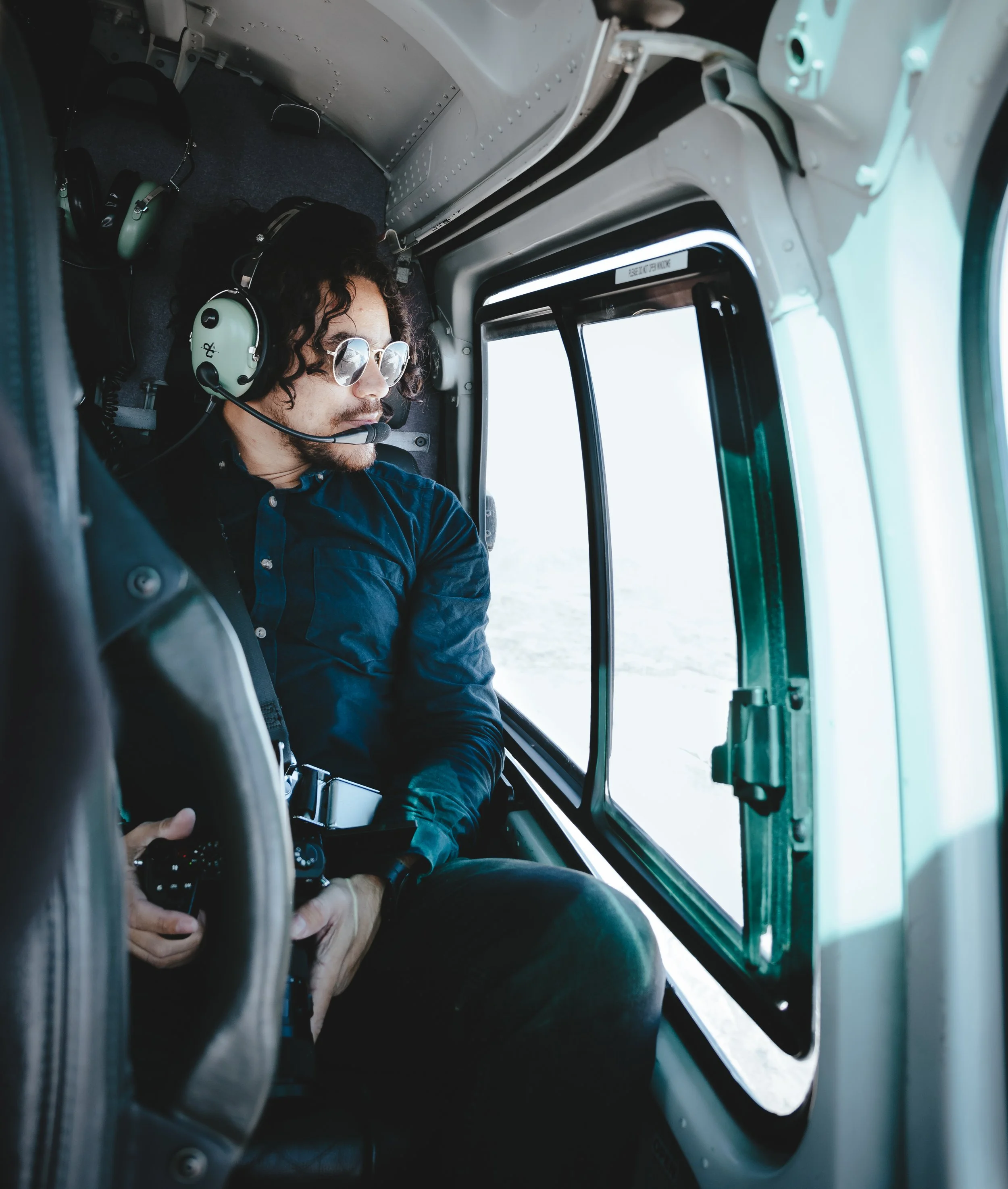 A man wearing sunglasses and a blue shirt is sitting in the cockpit of a helicopter. He is looking out the window, and a helicopter headset is nearby. The cockpit has metal and glass components, with a view of the sky and clouds outside.