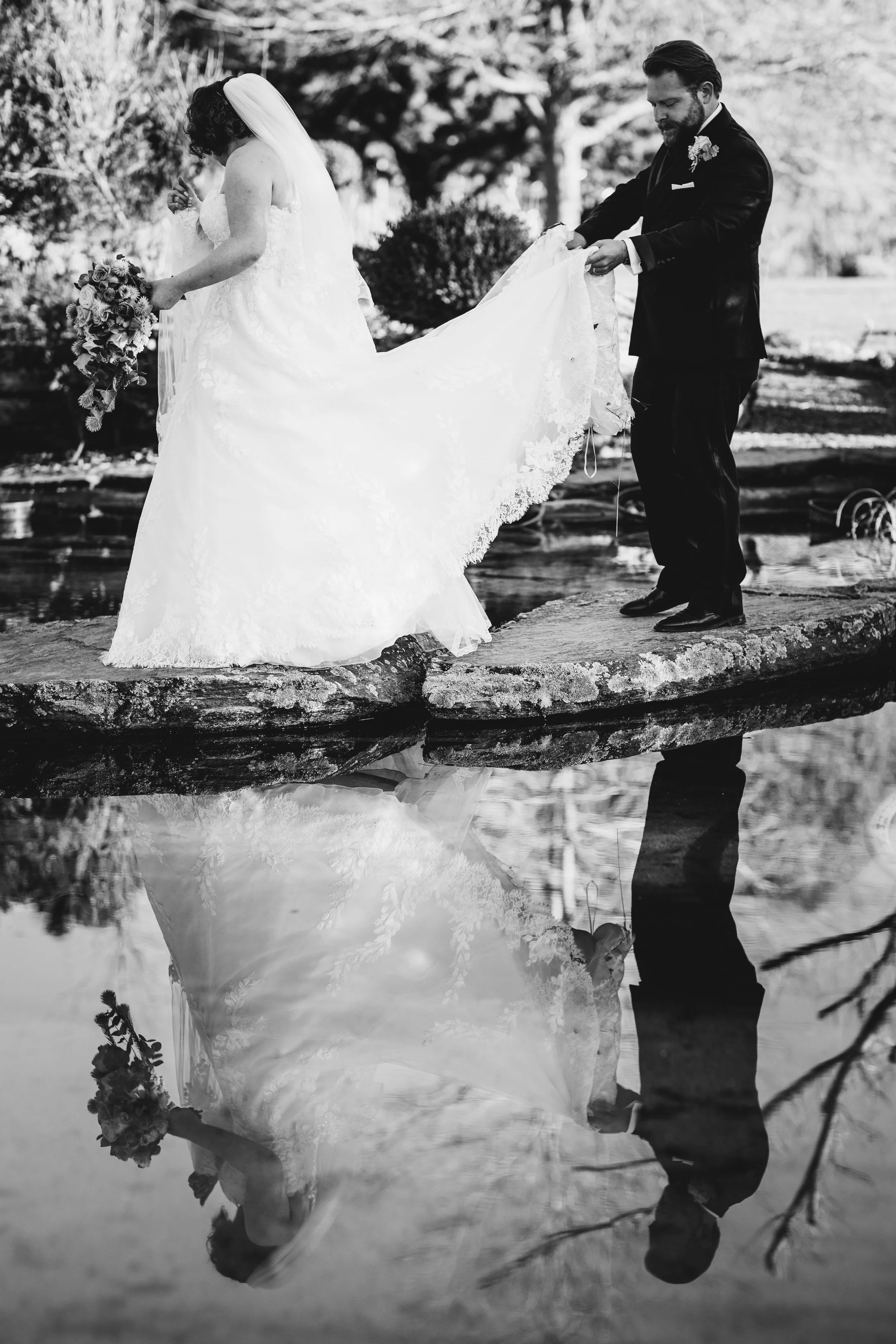A black and white photo of a bride and groom standing on a log by a river, with their reflection in the water. The bride is holding a bouquet and wearing a lace wedding gown, while the groom is in a dark suit with a boutonniere. They appear to be abo