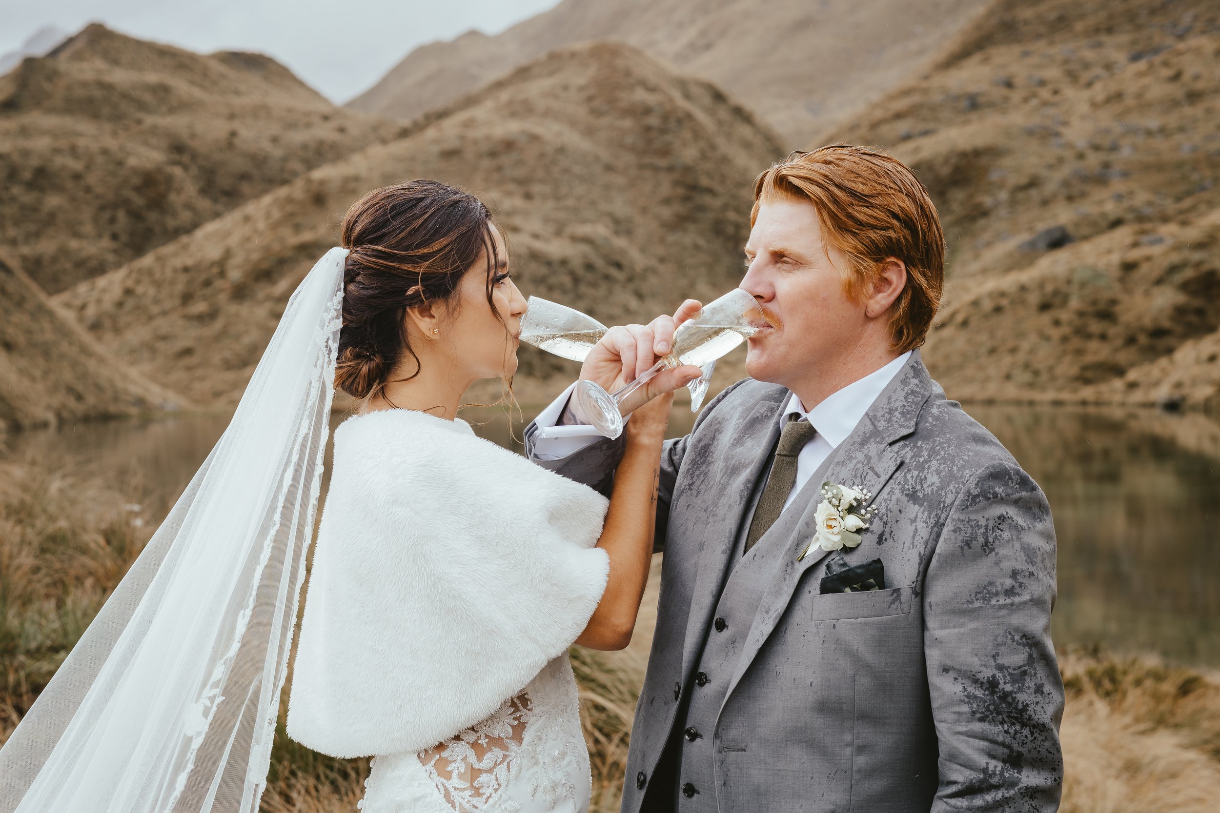 A bride and groom holding champagne glasses together, toasting during their outdoor wedding with a mountain landscape in the background.