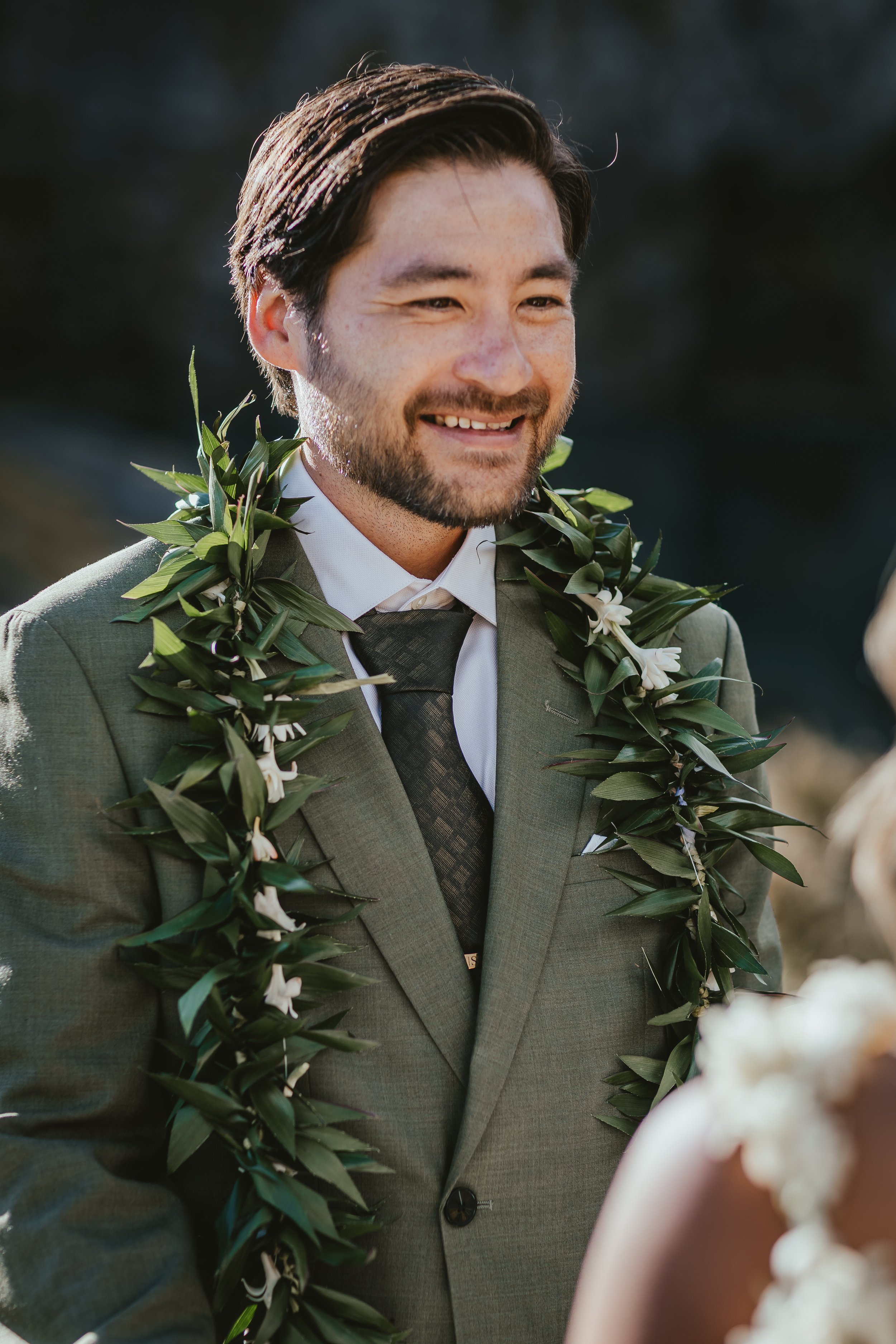 A smiling man with dark hair, beard, and mustache wearing a green suit with a white shirt and black tie, adorned with a lei made of green leaves and white flowers, standing outdoors against a blurred dark background.