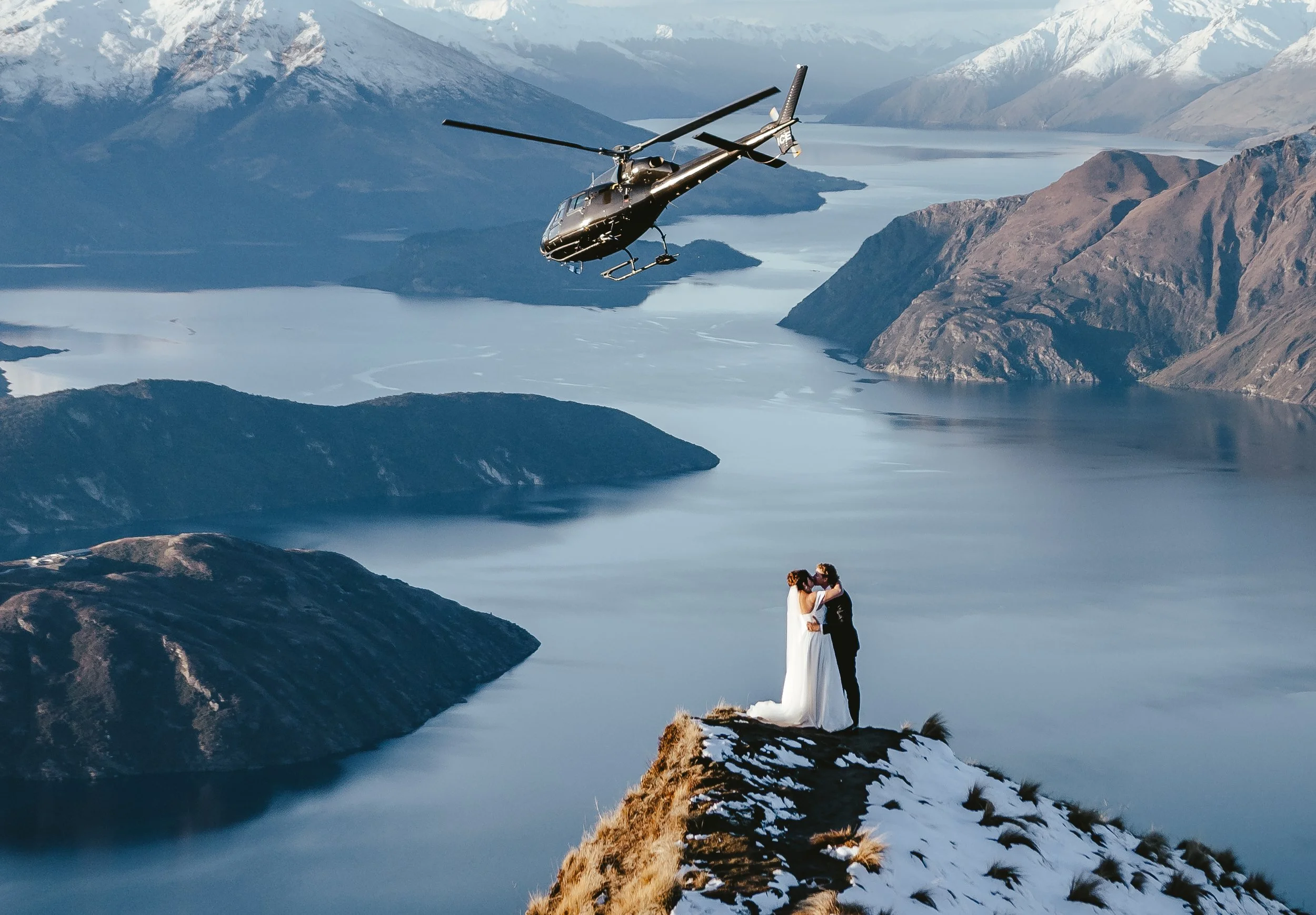 A couple dressed in wedding attire standing on a rocky ledge overlooking a lake surrounded by mountains, with a helicopter flying above them.