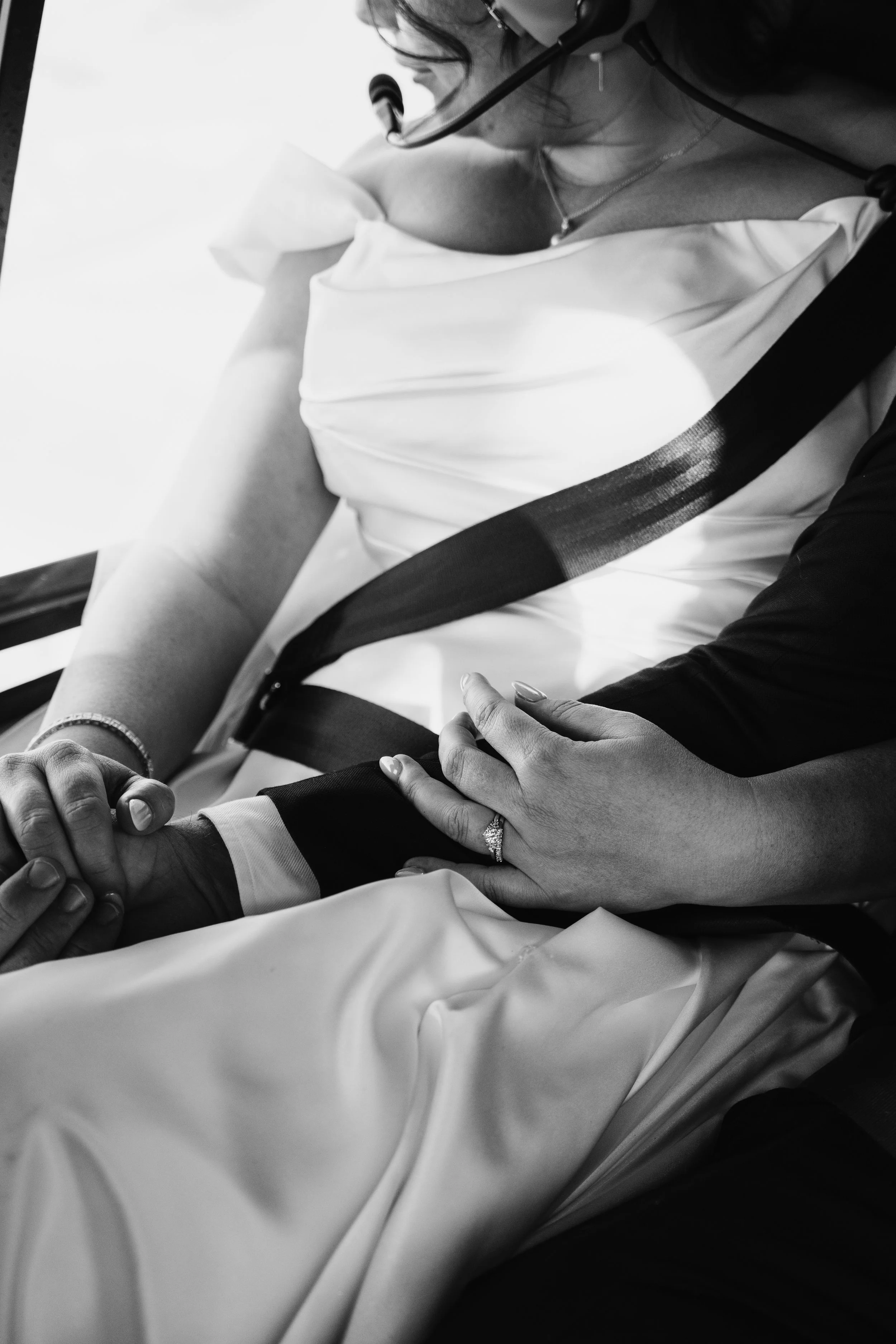 A black and white photo showing a bride and groom holding hands, with the bride wearing a white dress and engagement ring and the groom wearing a suit, attended in a tender moment.