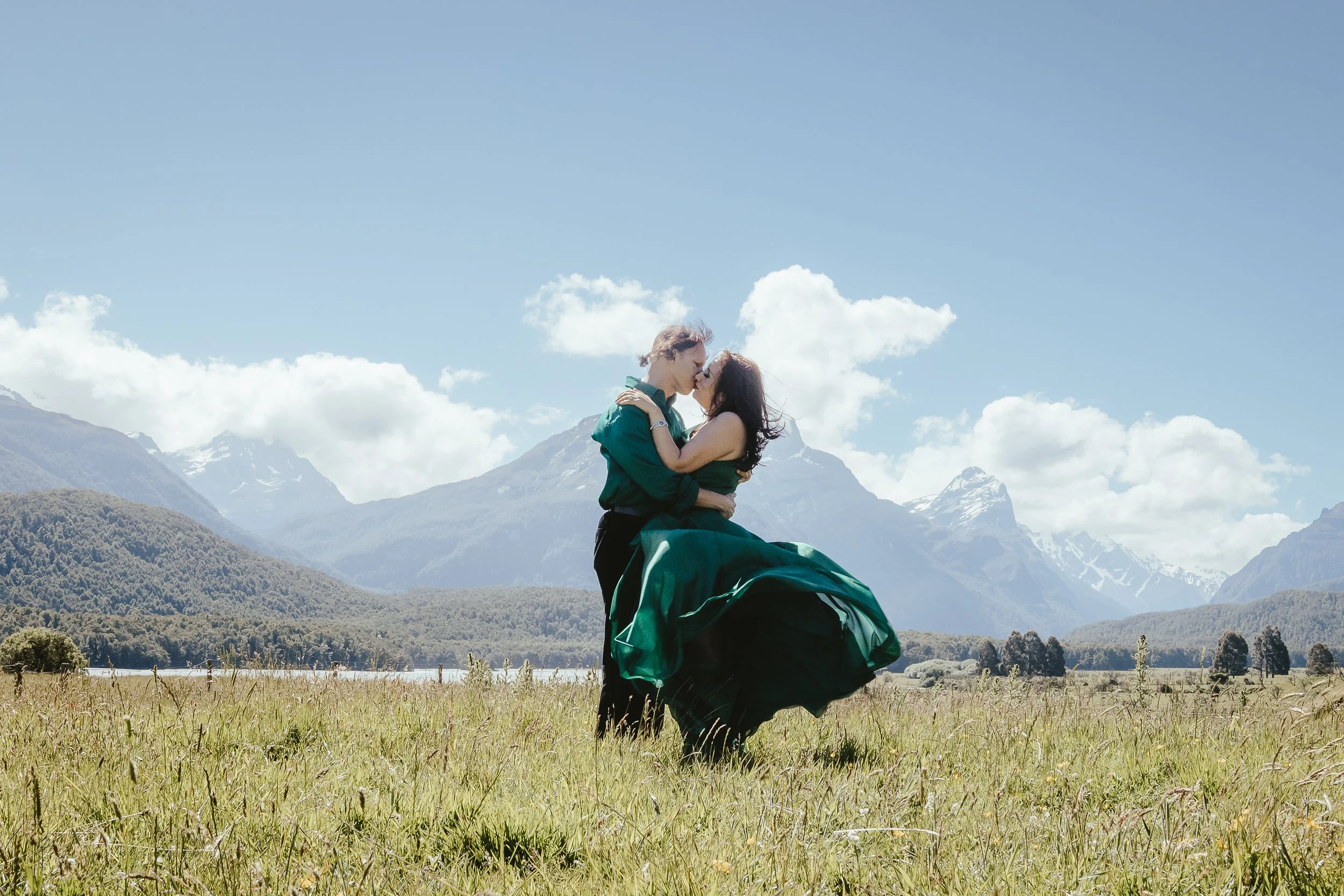 A couple kissing in a grassy field with mountains and blue sky in the background.