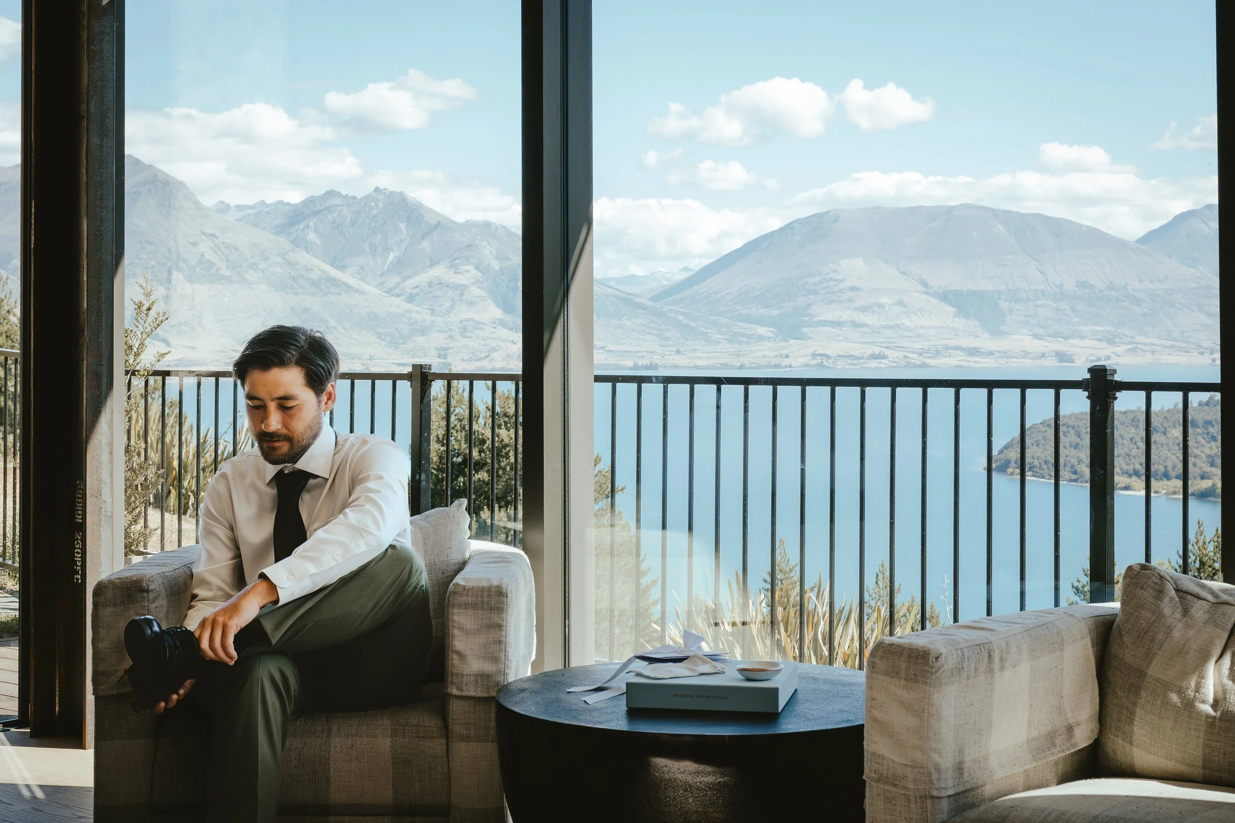 A man sits on a beige couch near large glass windows overlooking a mountain range and lake. He is dressed in a white shirt and black tie, looking down and adjusting his shoe. There is a small round table with papers and a box in front of him.
