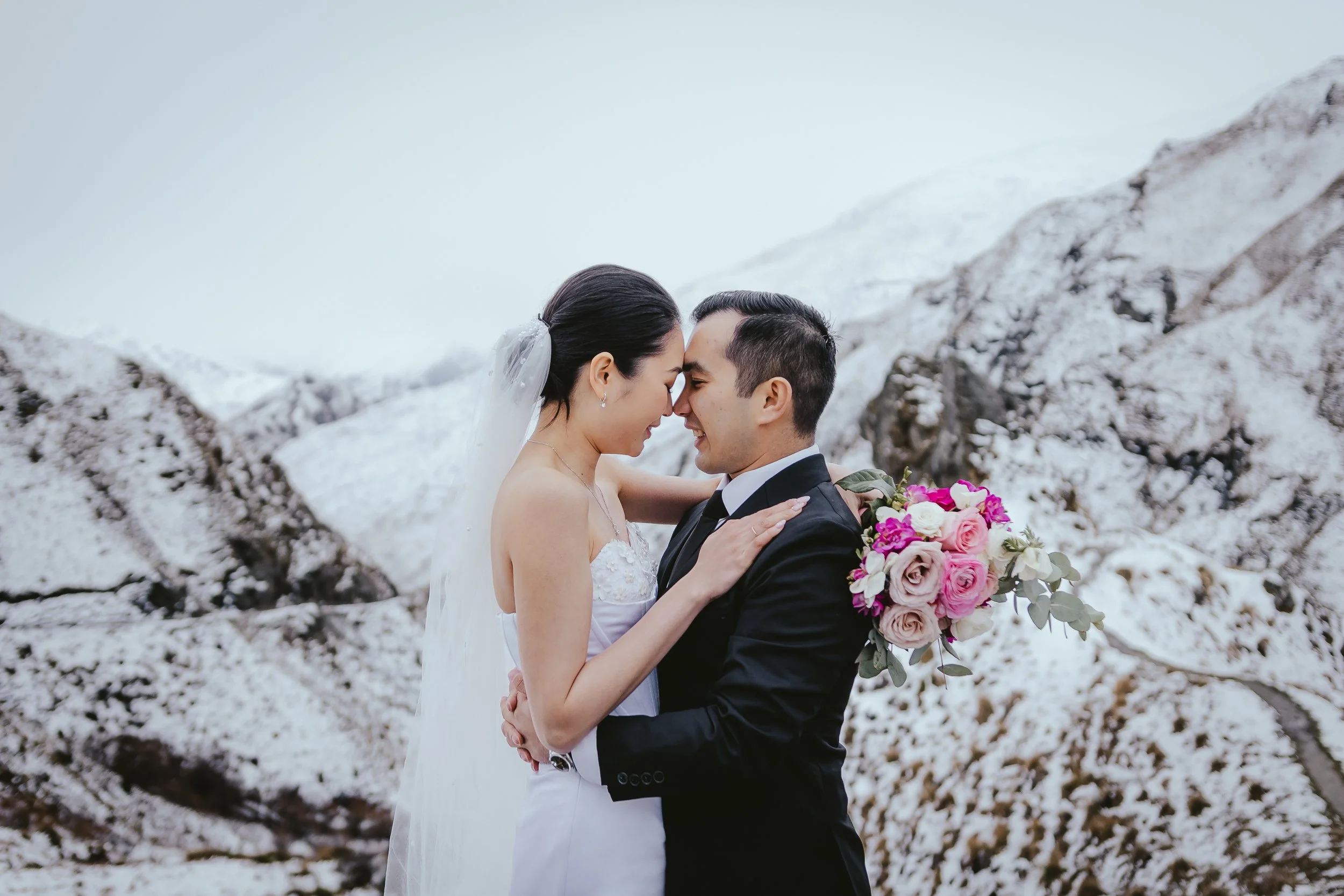 A bride and groom are embracing in front of snow-covered mountains during their wedding, with the bride holding a bouquet of pink and white roses.