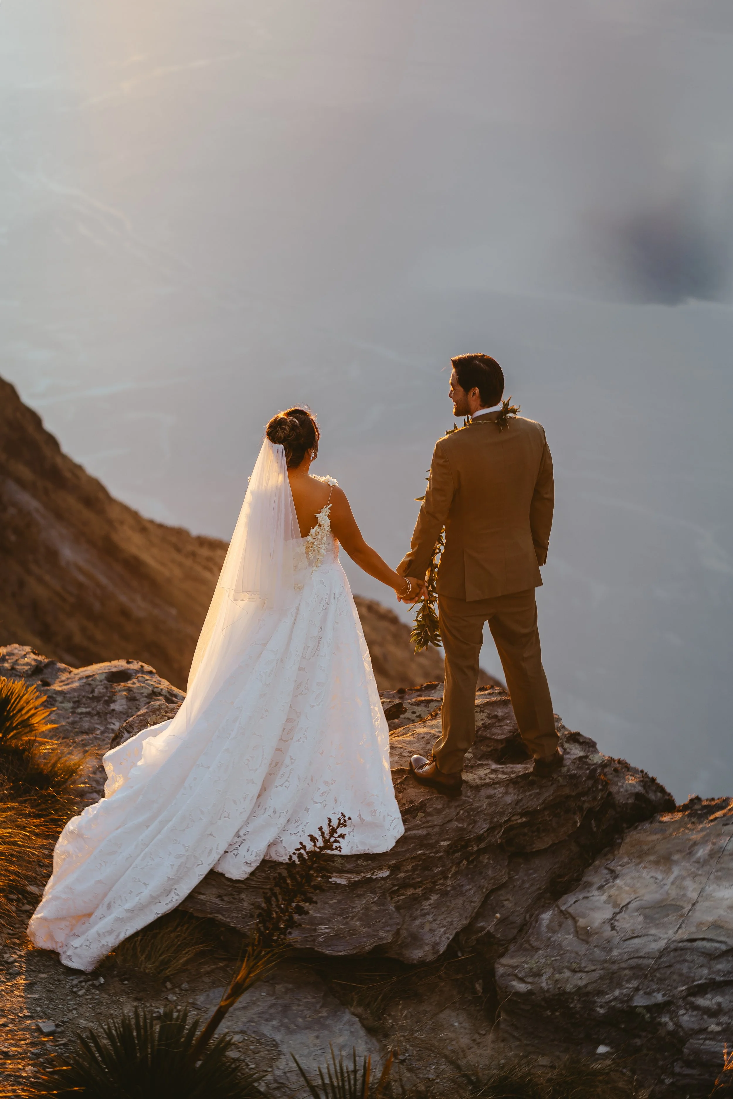 A bride and groom standing hand in hand on a rocky ledge at sunset, overlooking a cloudy sky and mountains in the distance.