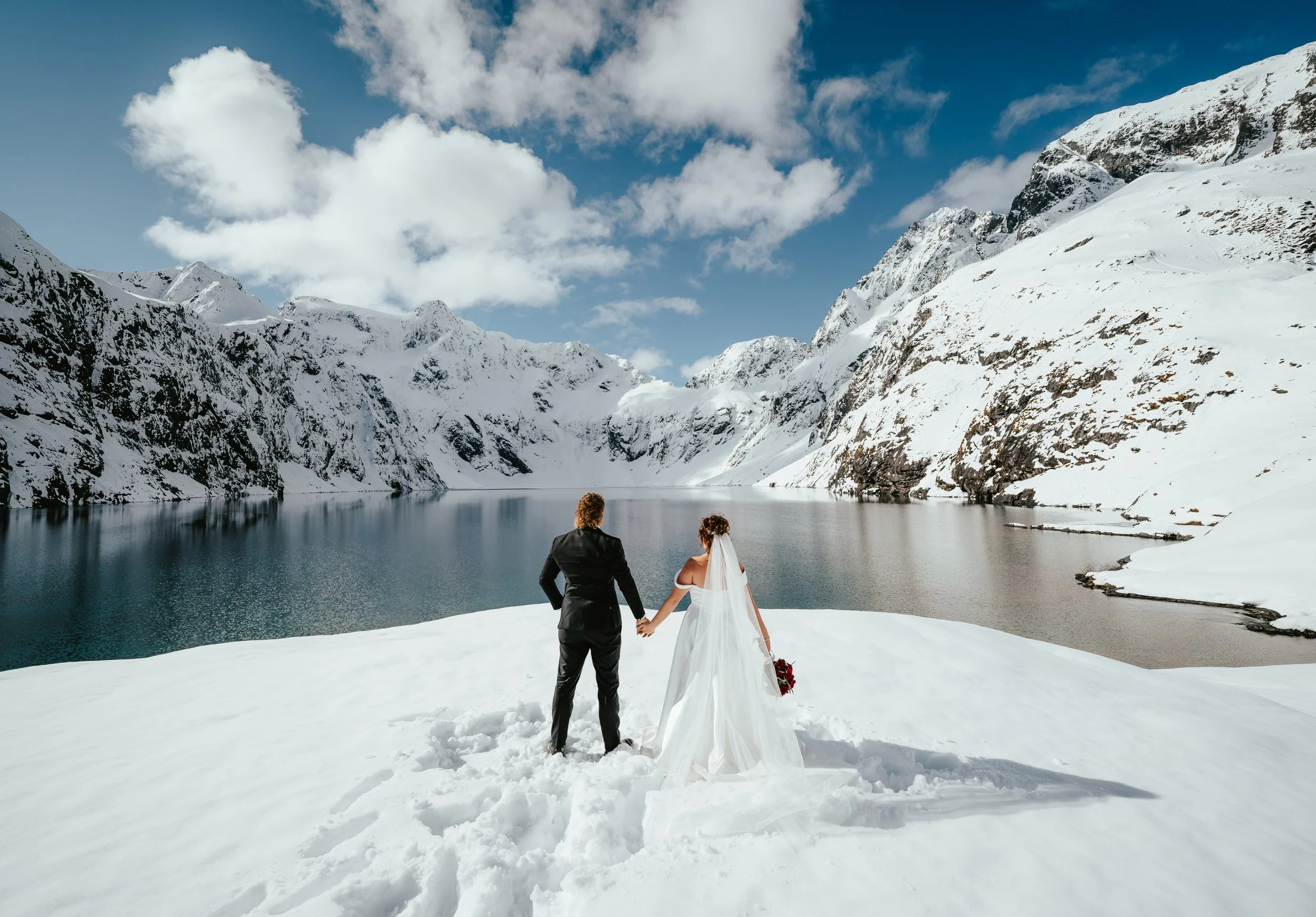 A bride and groom holding hands in snow-covered terrain near a fjord, with snow-capped mountains and partly cloudy sky in the background.