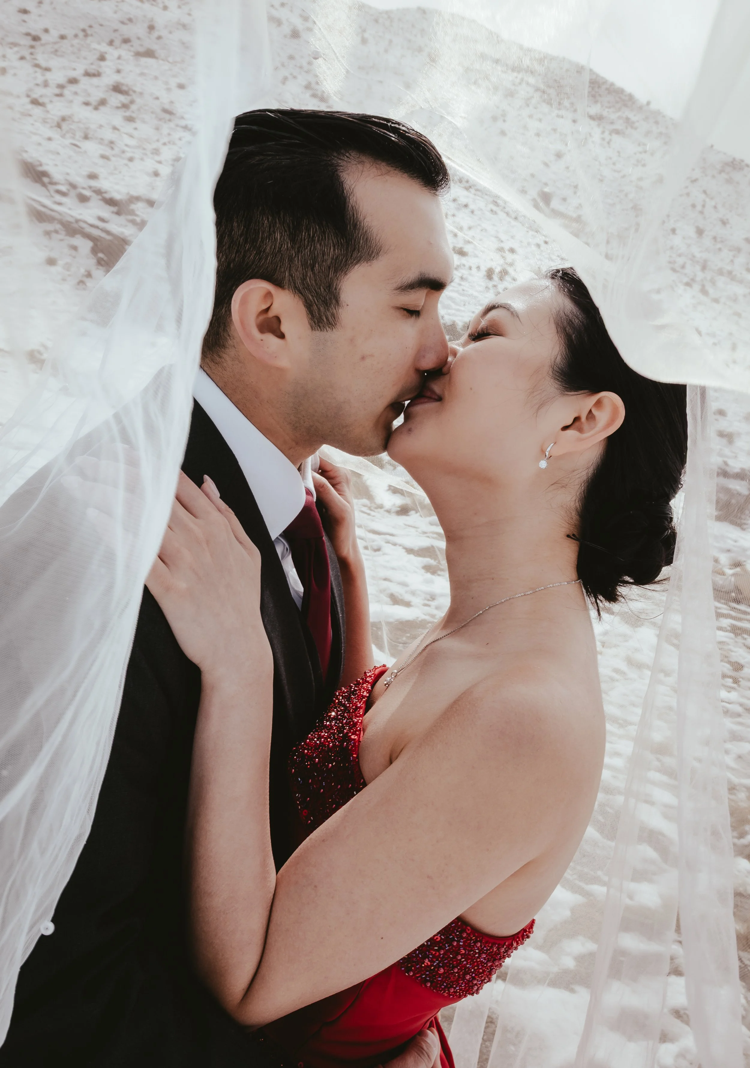 A couple kissing under a wedding veil on a sandy beach.