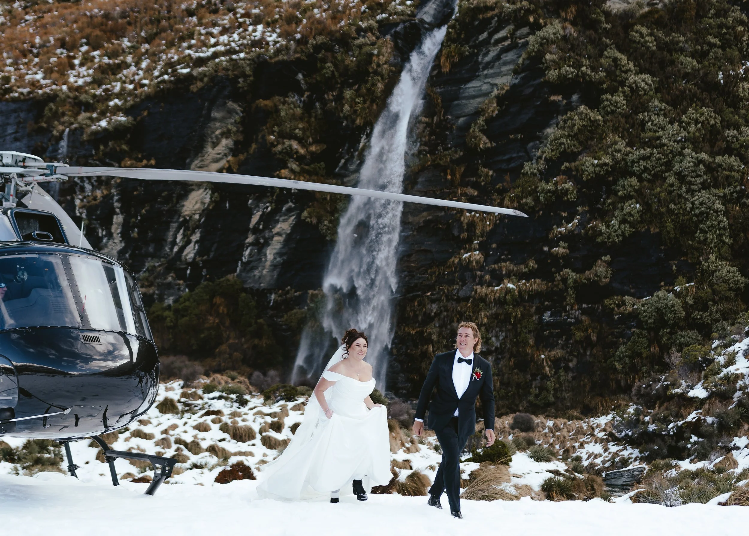 Couple walking away from a helicopter after landing at a frozen waterfall site for their private heli elopement.