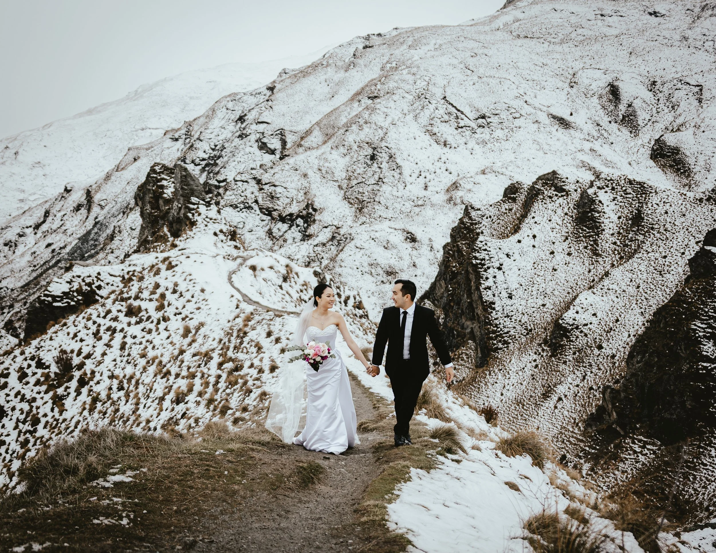A couple dressed in wedding attire walking hand in hand on a snowy mountain trail, with snow-covered mountains in the background.