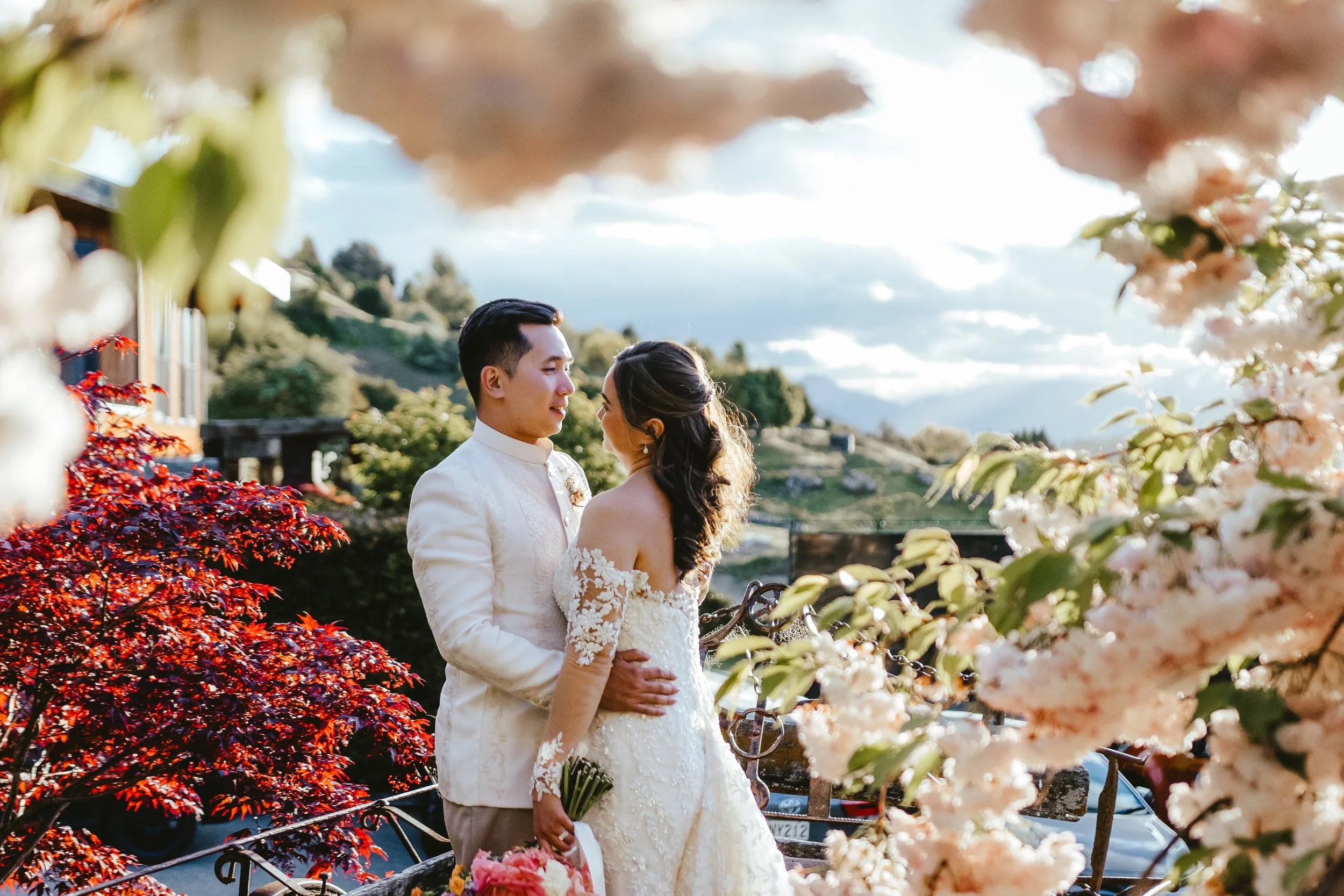 A couple in wedding attire standing close together outdoors, surrounded by blooming pink and red flowers with mountains and a cloudy sky in the background.