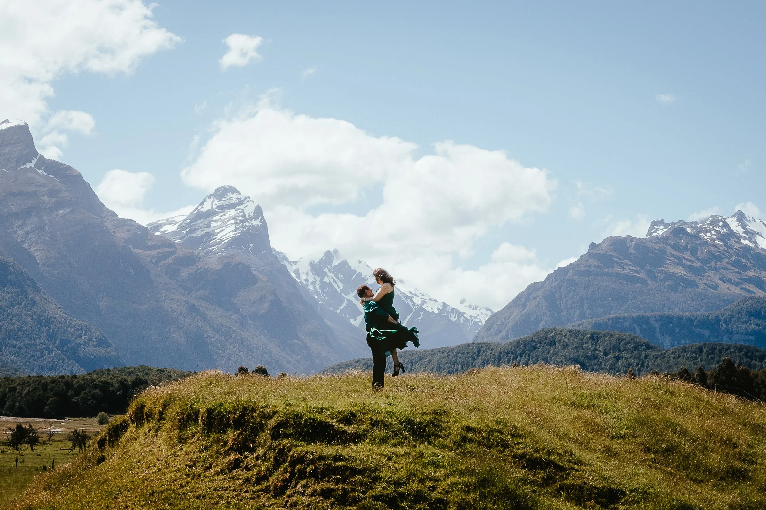 A person carrying a child on their shoulders in a grassy field with tall snow-capped mountains and partly cloudy sky in the background.