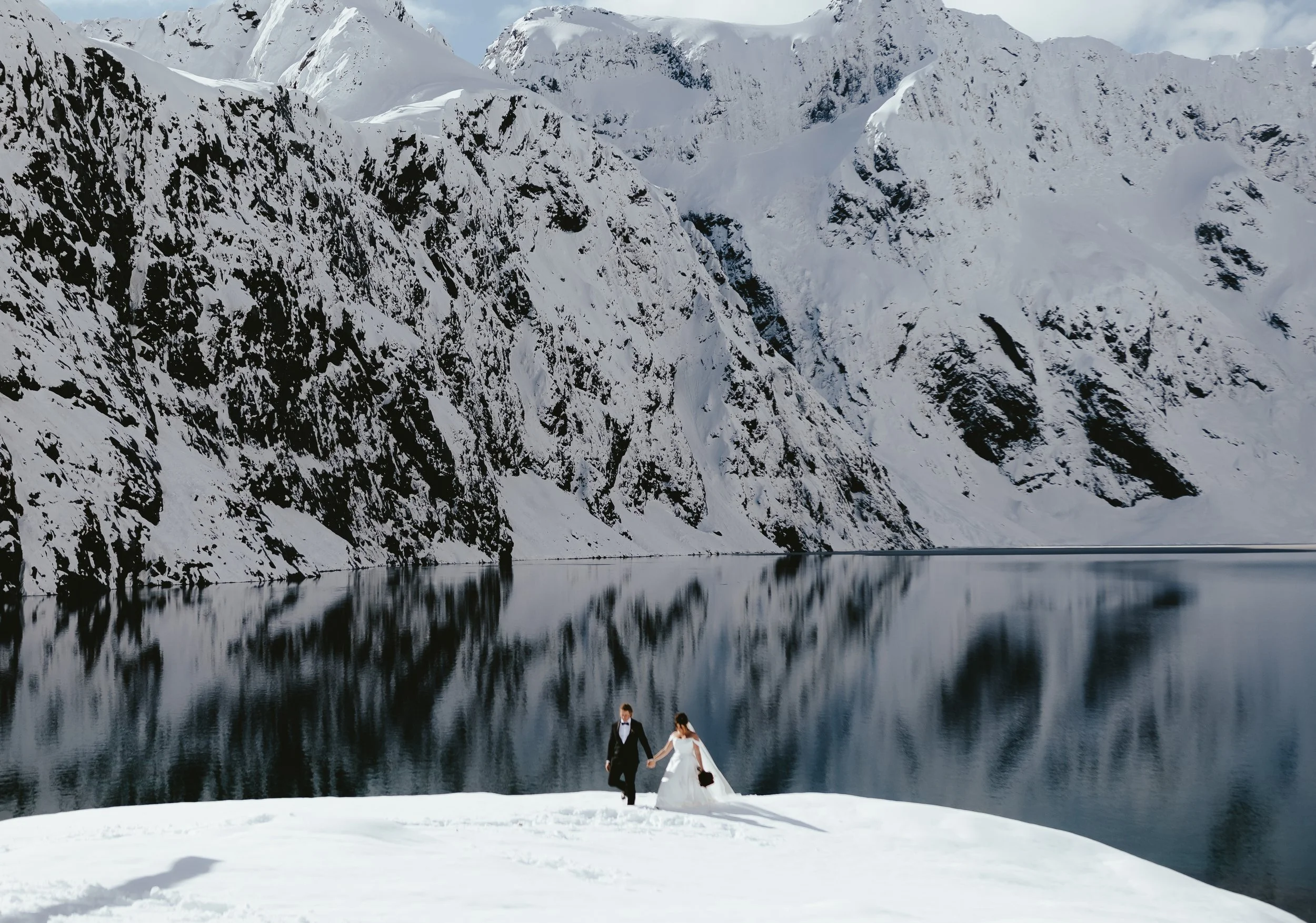 A bride and groom holding hands and walking along a snowy lakeshore, with snow-covered mountains and their reflection in the calm water behind them.