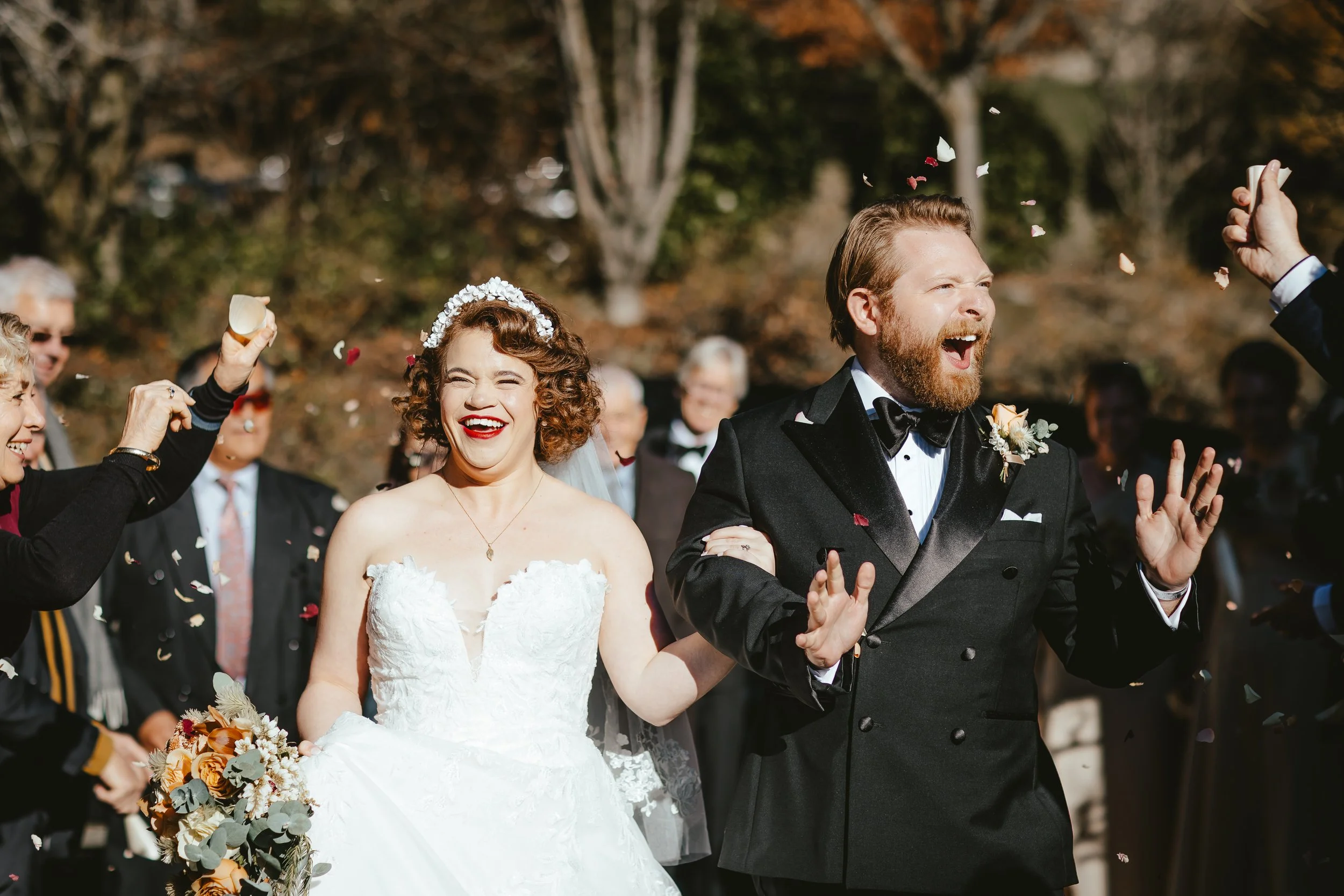 A bride and groom celebrating their wedding outdoors with guests throwing flower petals. The bride wears a white wedding dress and veil, holding a bouquet, and has curly hair with a floral headpiece. The groom is in a black tuxedo with a bow tie and 