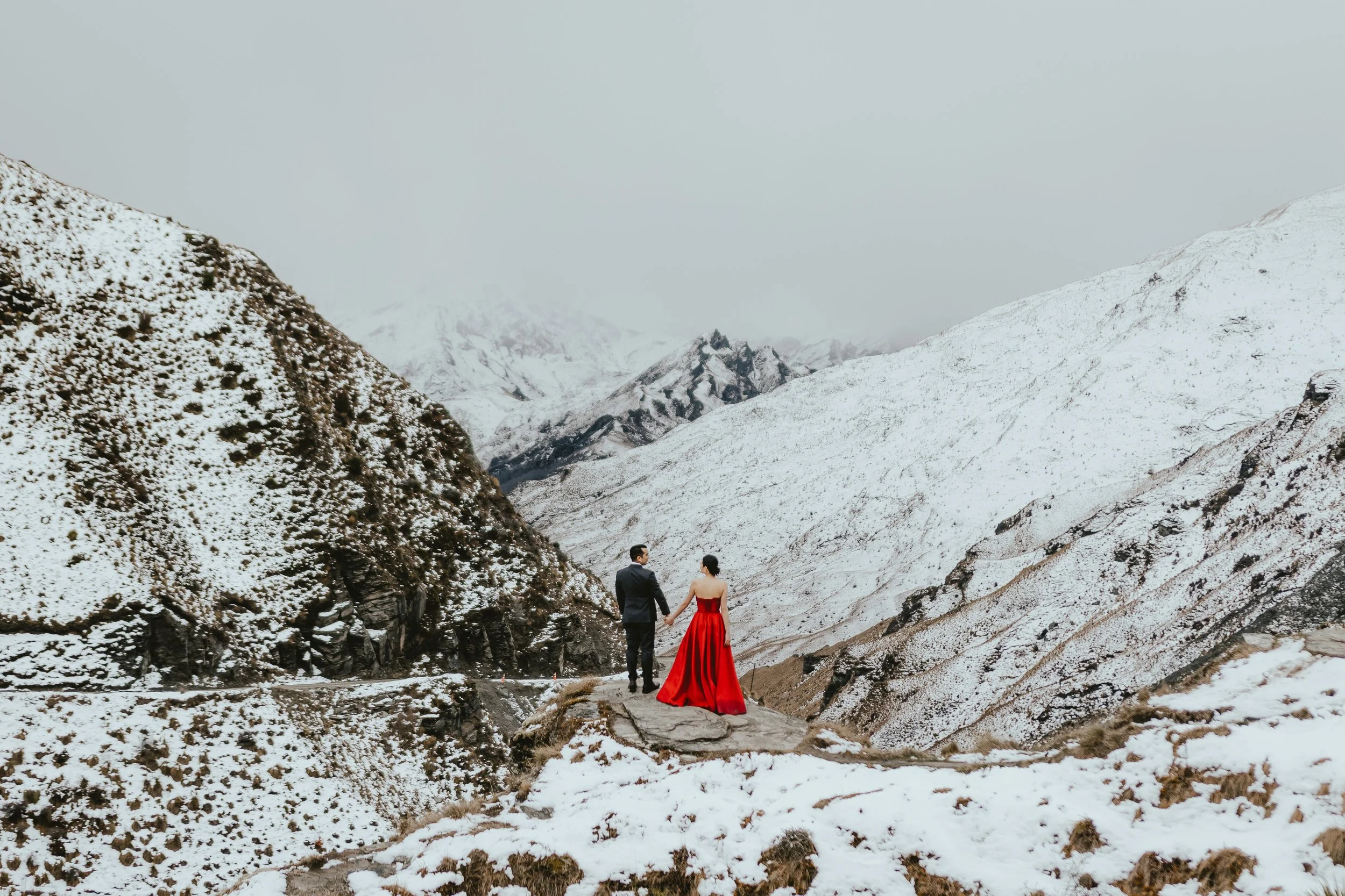 A couple standing hand in hand in a snow-covered mountainous landscape, with the woman wearing a long red dress and the man in a dark suit.