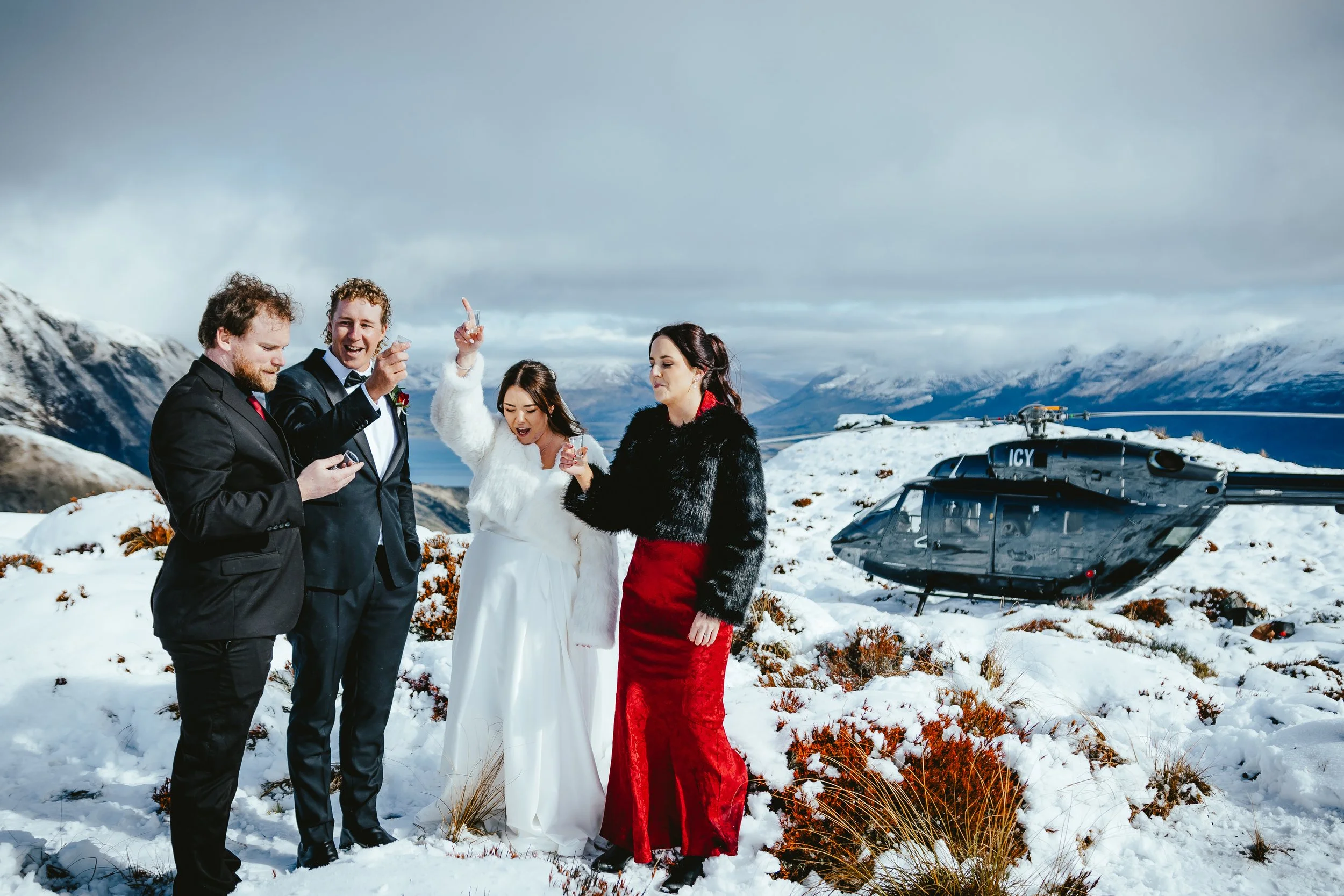 Group of five people dressed in formal attire standing outdoors in a snowy mountainous area with a helicopter nearby, during daytime, celebrating a special occasion.