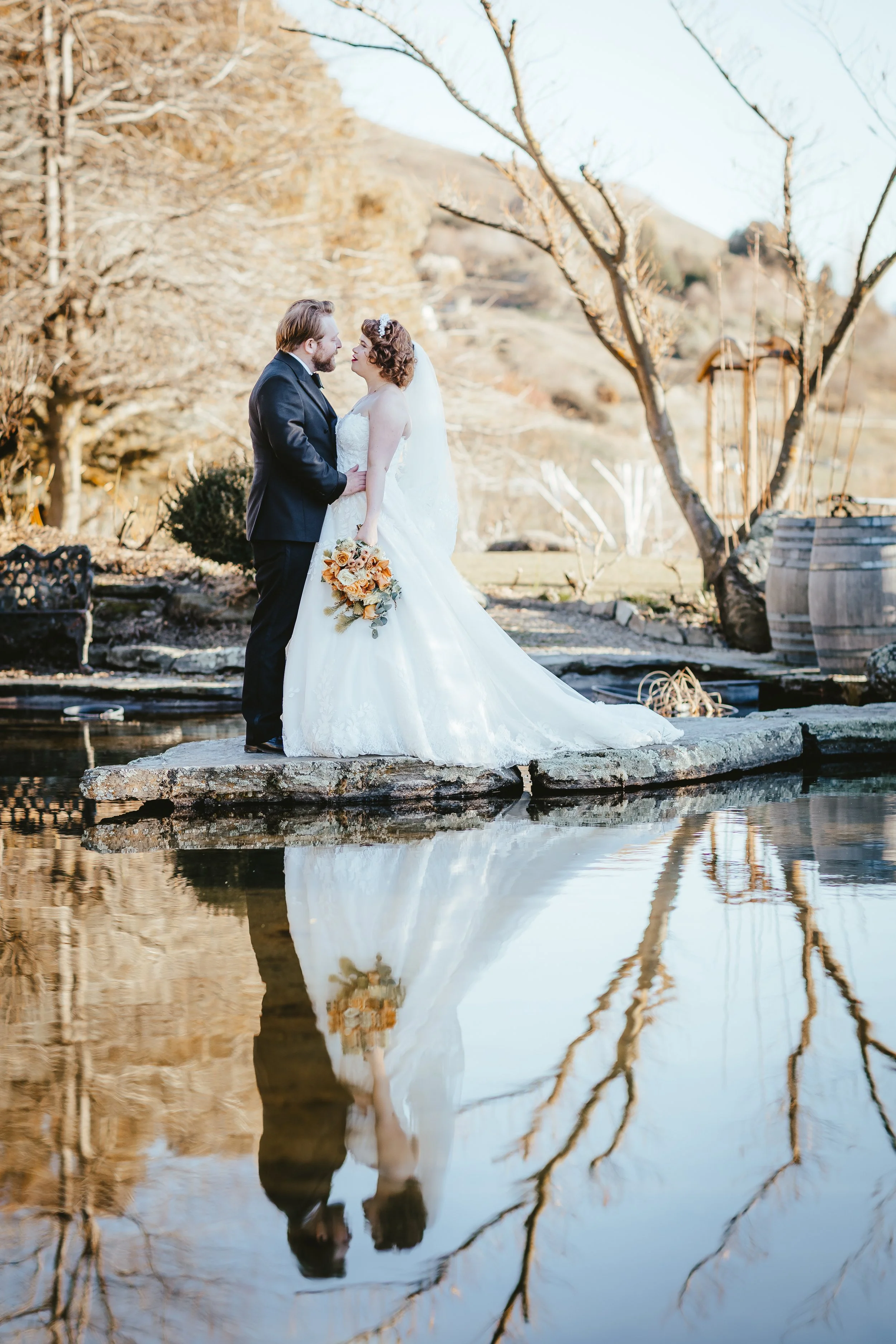 A bride and groom are standing on a stone platform by the water, gazing into each other's eyes. The bride is holding a bouquet of orange and cream flowers. The background features bare trees and a rustic barrel, with their reflections visible in the 
