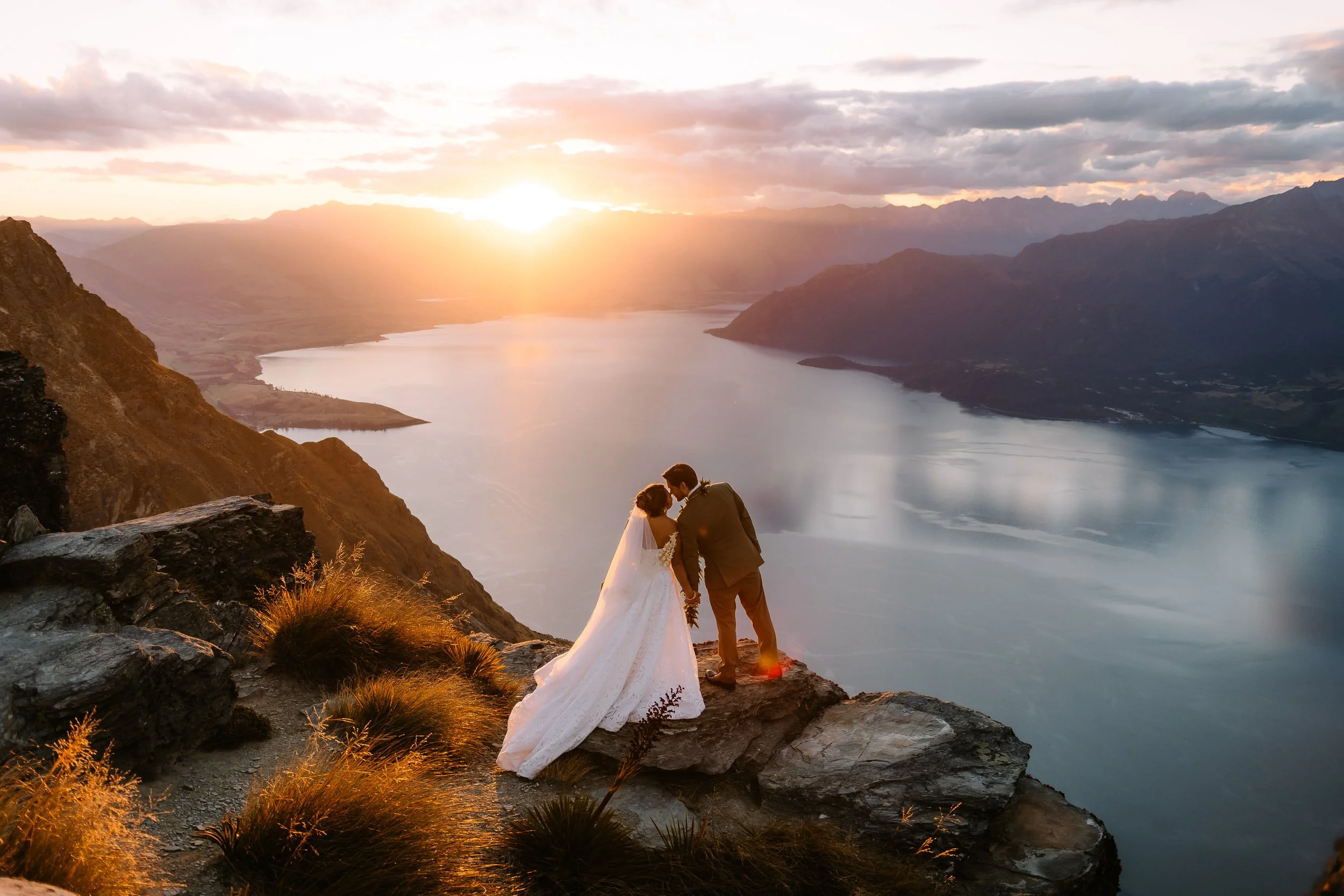 A bride and groom standing on a rocky ledge overlooking a large lake or river with mountains in the background during sunset. The bride is wearing a white wedding gown and the groom is in a dark suit. They are holding hands and leaning in towards each other.