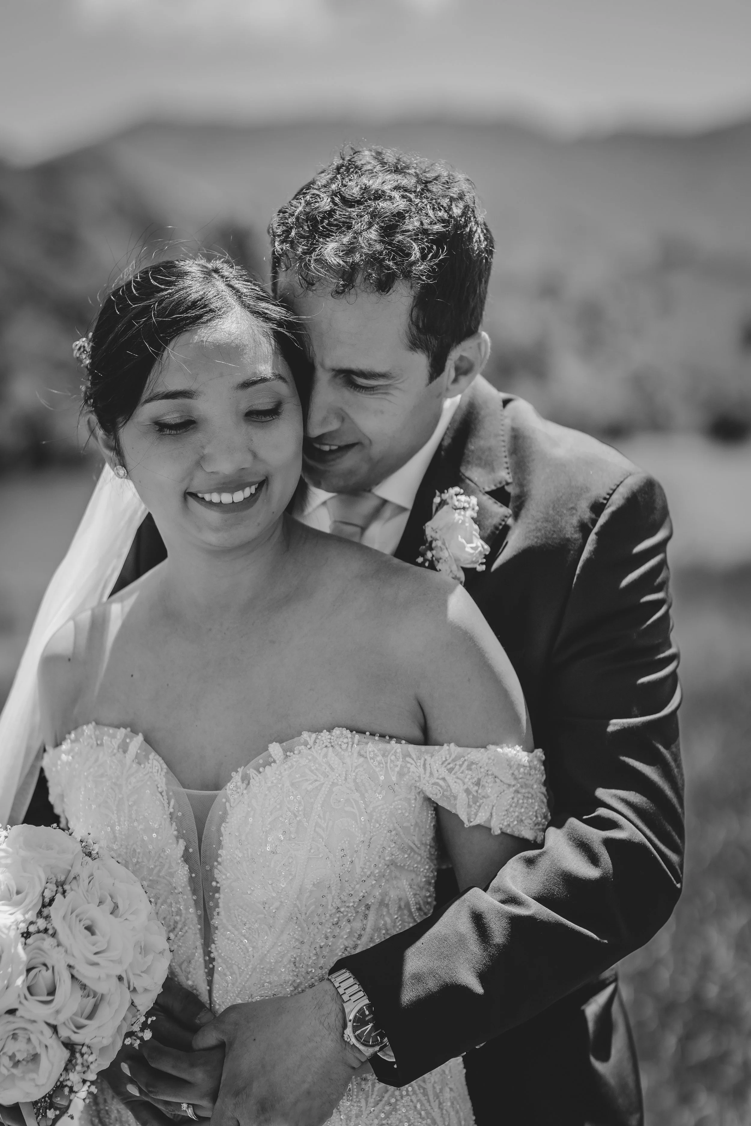 A black and white photo of a smiling bride and groom embracing outdoors. The bride wears an off-the-shoulder lace wedding dress and holds a bouquet of flowers. The groom is in a suit with a boutonniere, wearing a wristwatch. They are close together, 