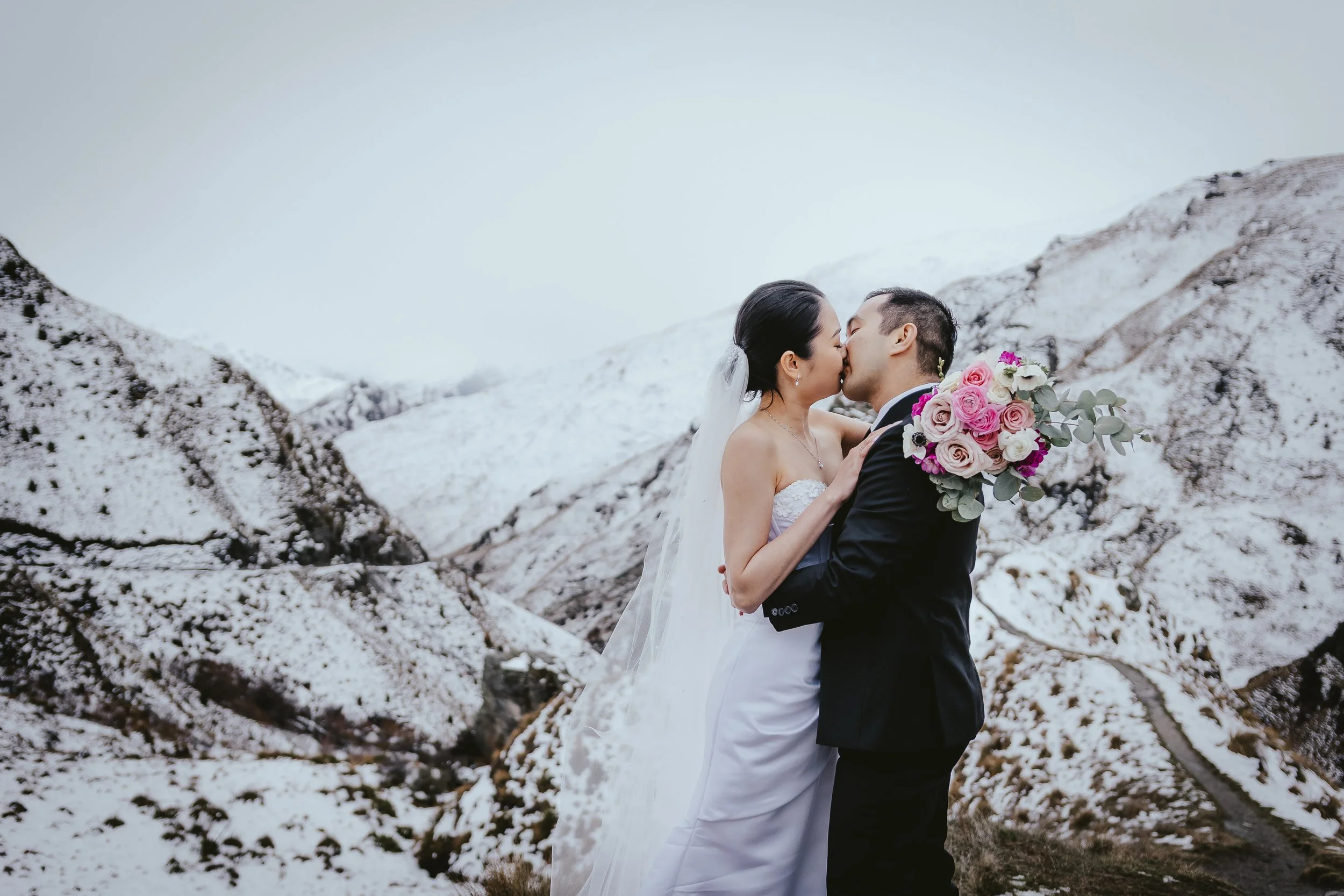 A bride and groom kissing in a snowy mountainous landscape, with the bride holding a bouquet of pink and white roses.