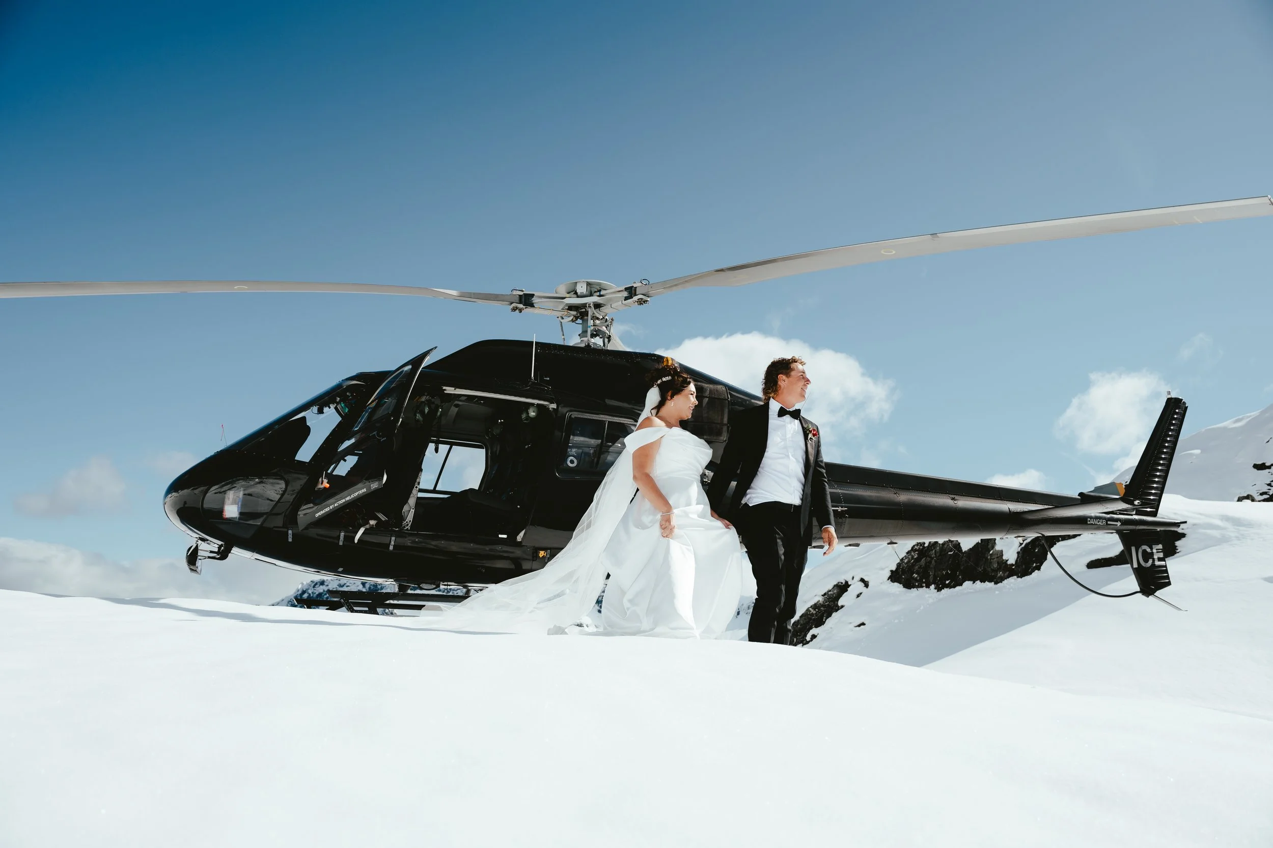 Bride and groom in wedding attire standing next to a black helicopter on snowy mountain landscape with clear blue sky.
