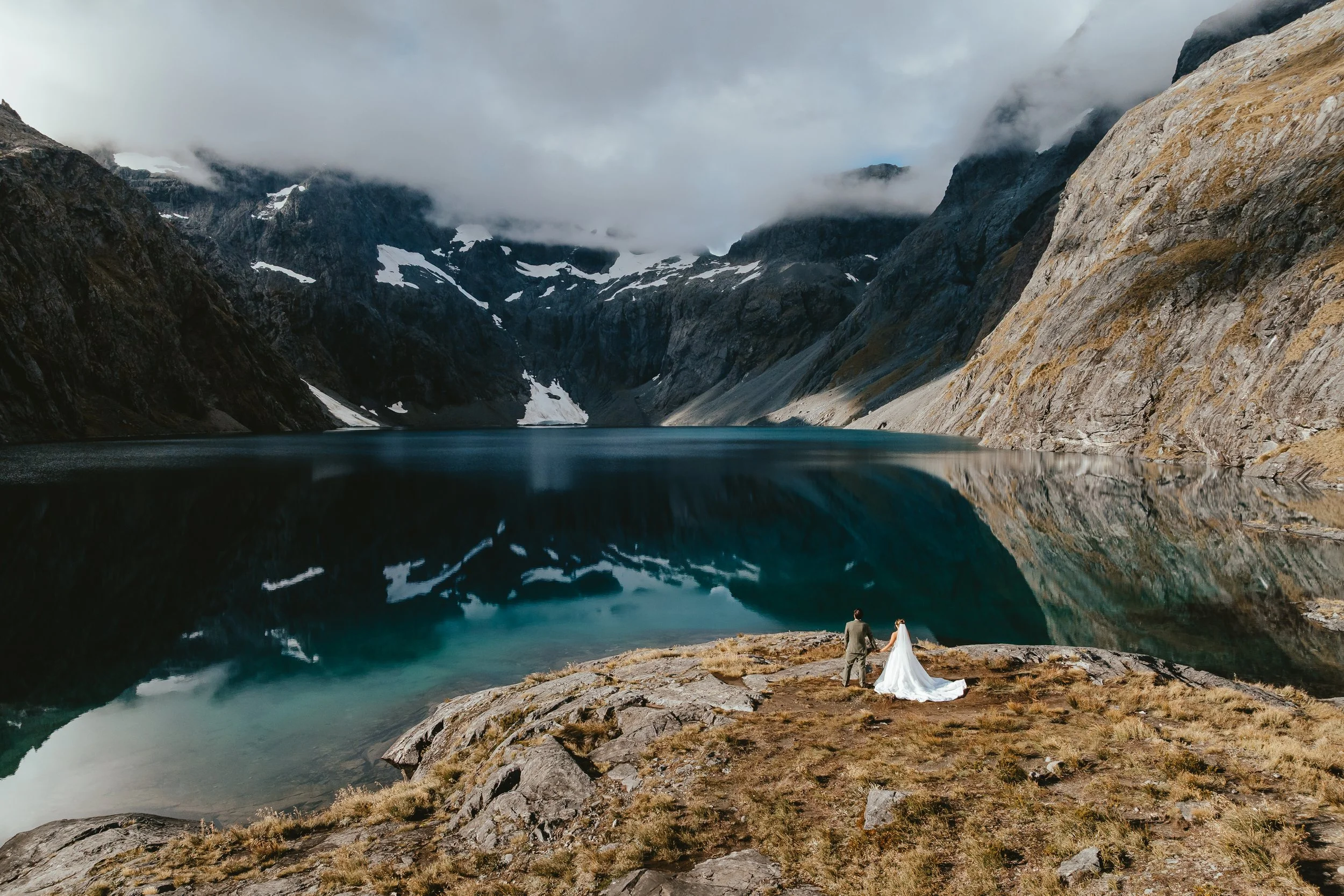 A couple dressed in wedding attire standing on rocks near a mountain lake, with steep rocky mountains and clouds in the background.