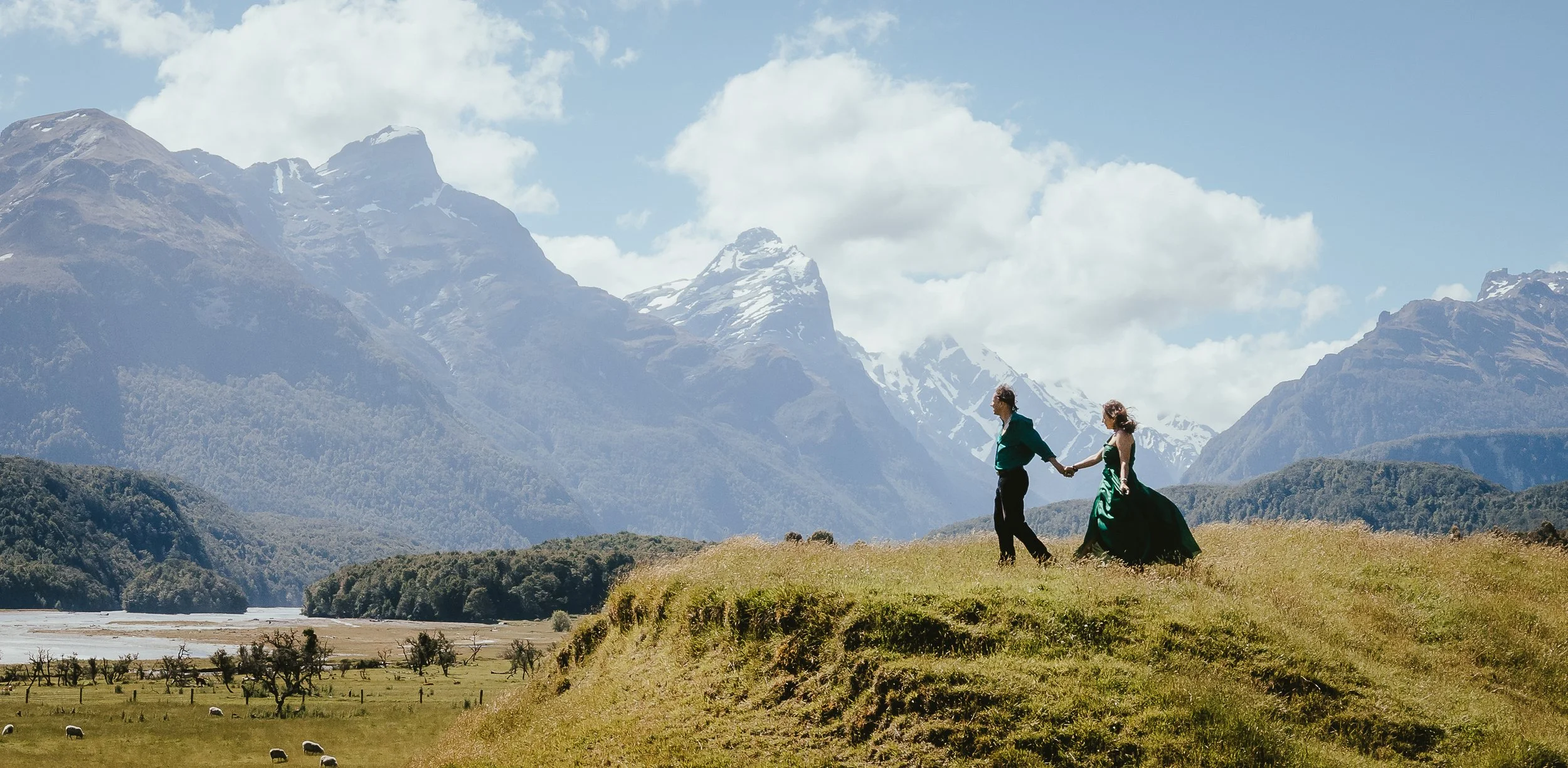 A couple holding hands, walking on a grassy hill with mountains in the background under a partly cloudy sky.