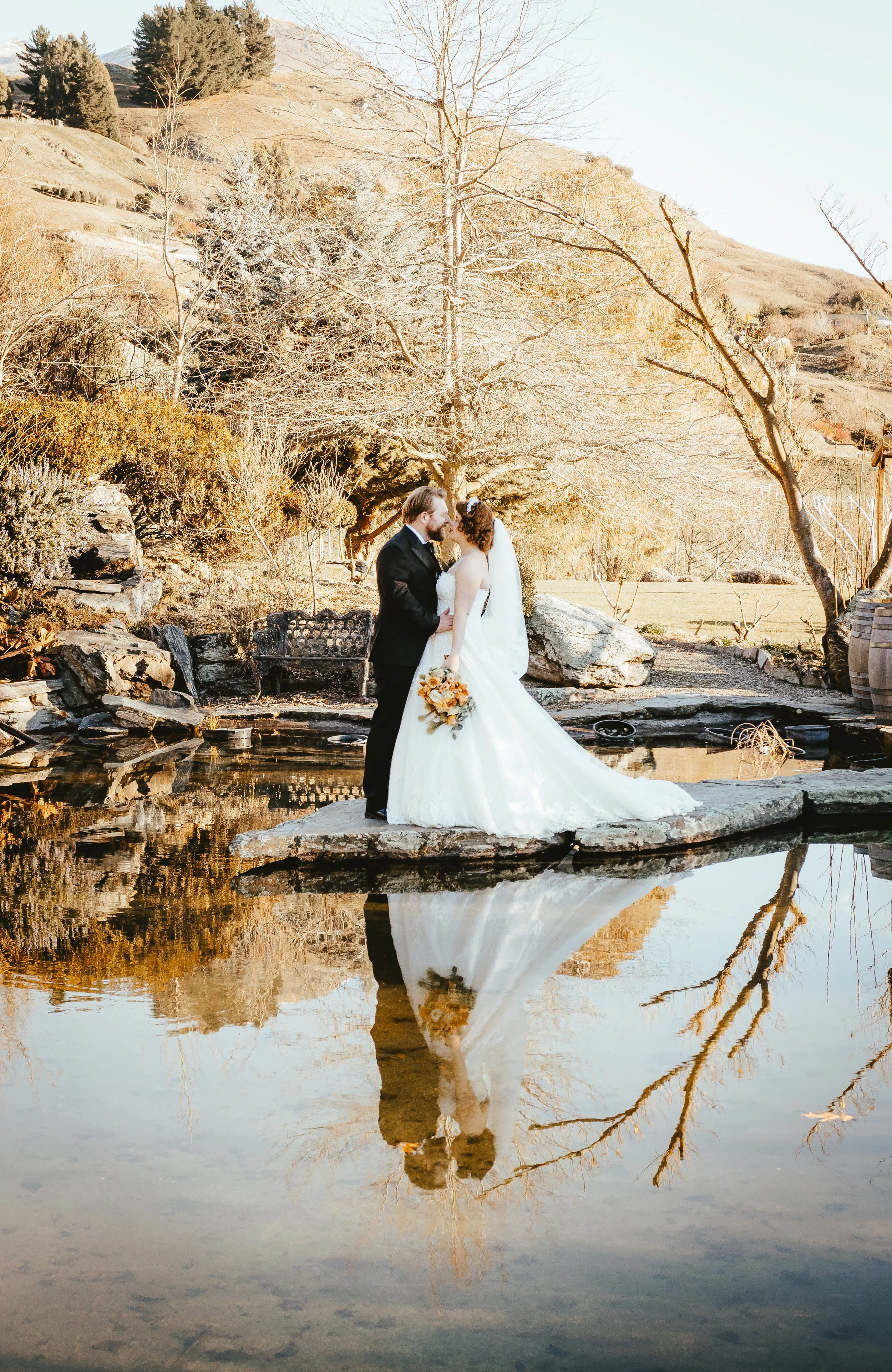 A bride and groom kissing on a rock by a pond, with their reflection visible in the water. The background features leafless trees and hills, suggesting a fall or winter setting.