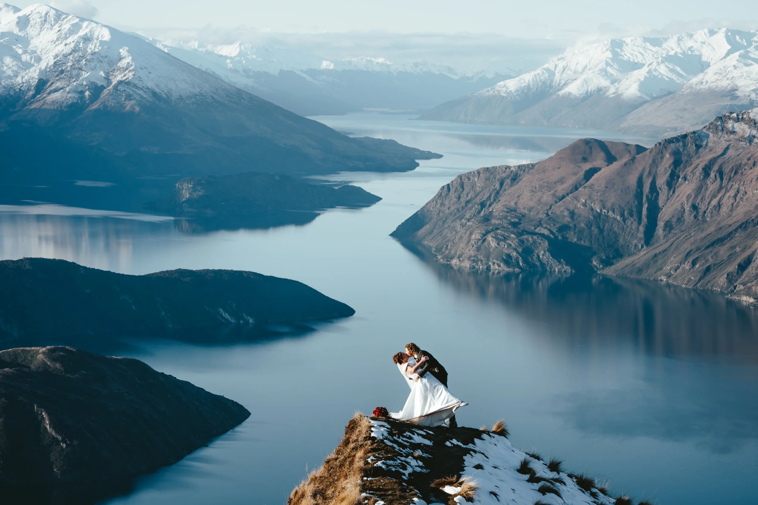A bride and groom kissing on a mountain overlooking a lake and snow-capped mountains.