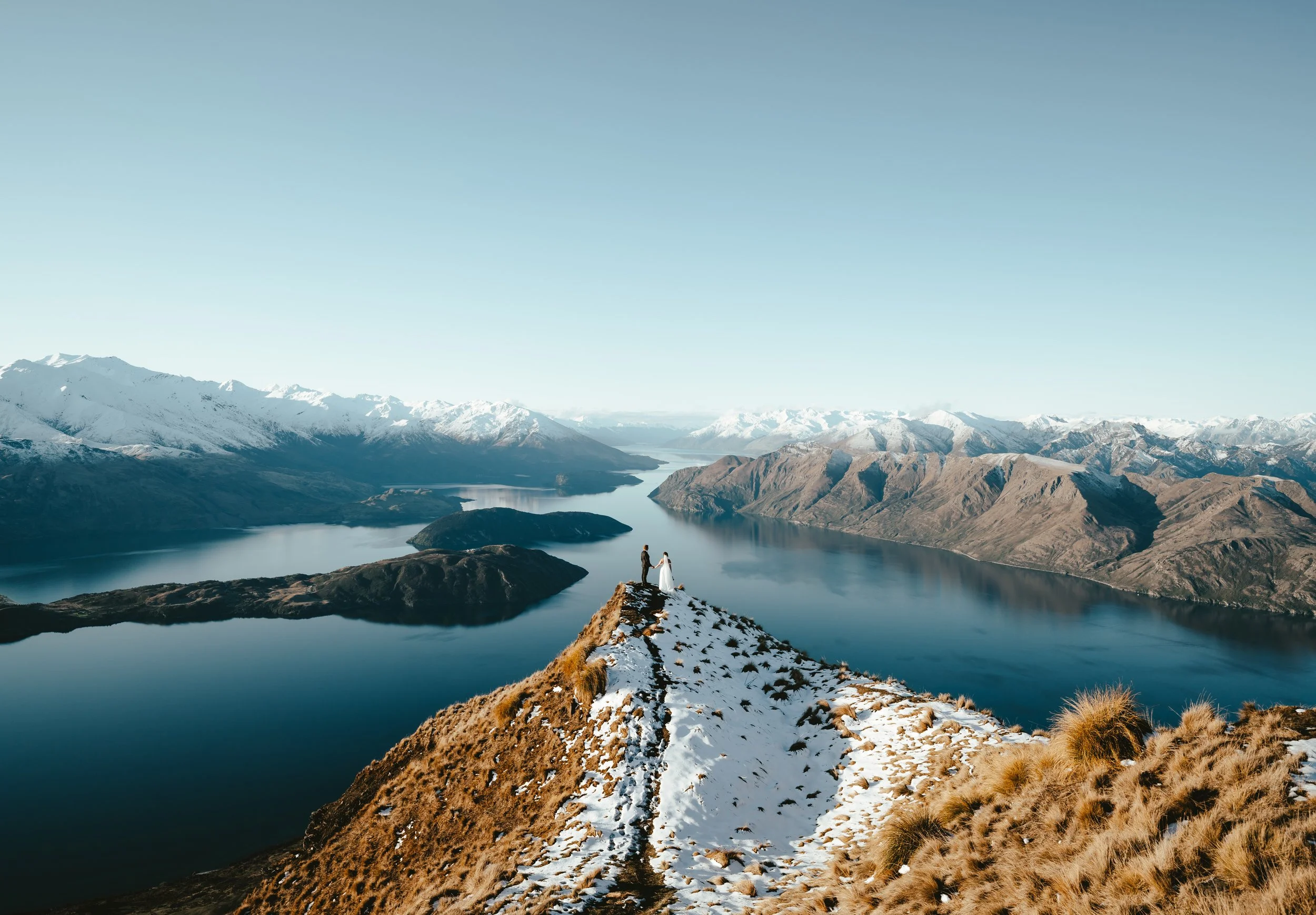 A couple holding hands standing on a narrow trail atop a mountain ridge with snow patches, overlooking a large body of water surrounded by mountain ranges with snow-capped peaks under a clear blue sky.