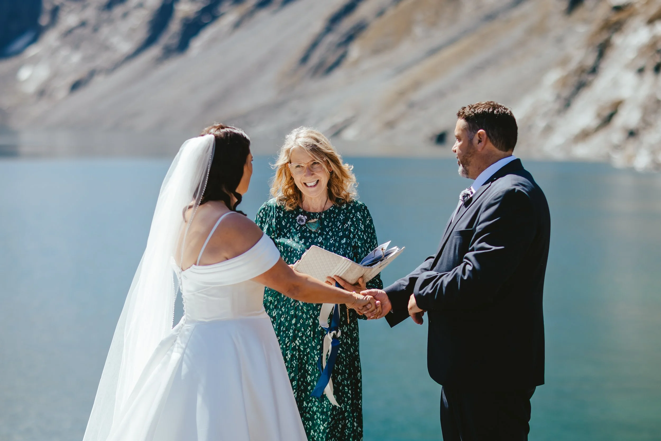 Bride and groom holding hands during wedding ceremony outdoors near a body of water, officiant smiling and reading from a book between them, mountainous landscape in background.