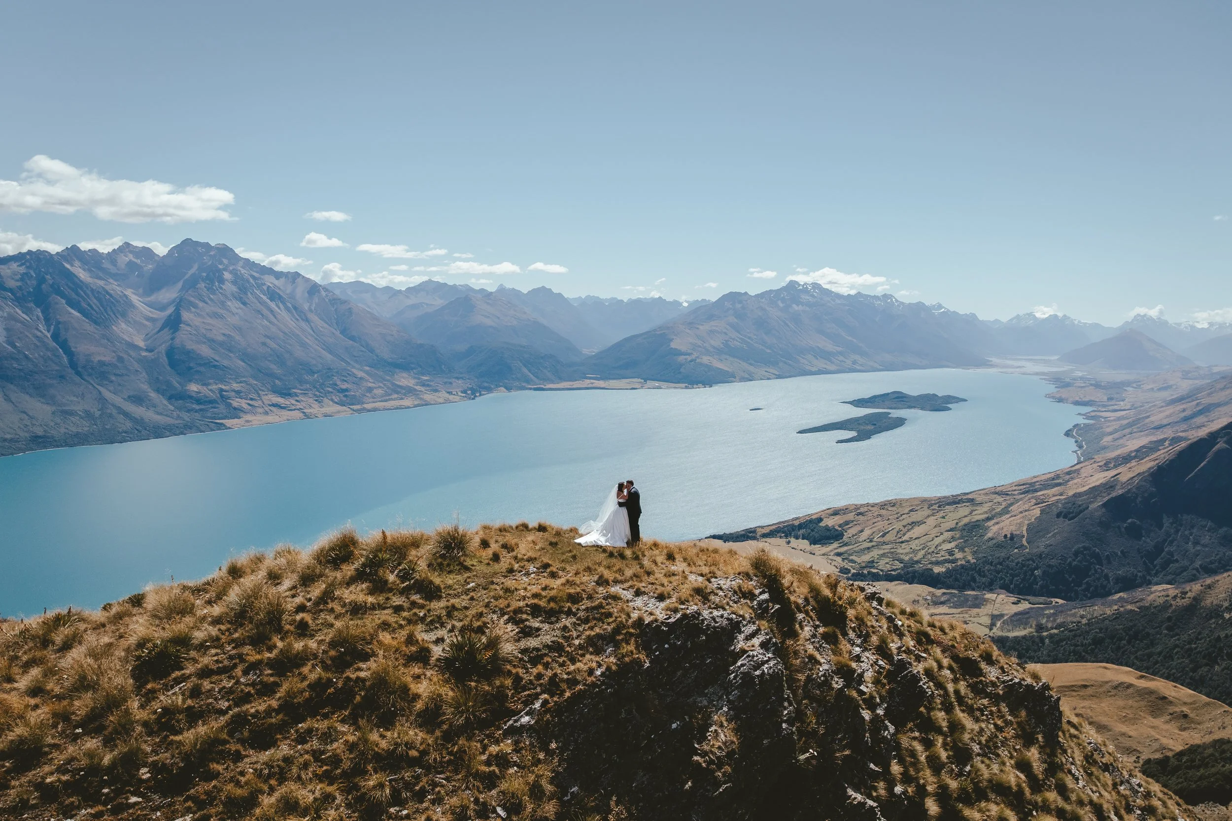 A bride and groom sharing a kiss on a hilltop overlooking a large lake, mountains, and a clear blue sky.