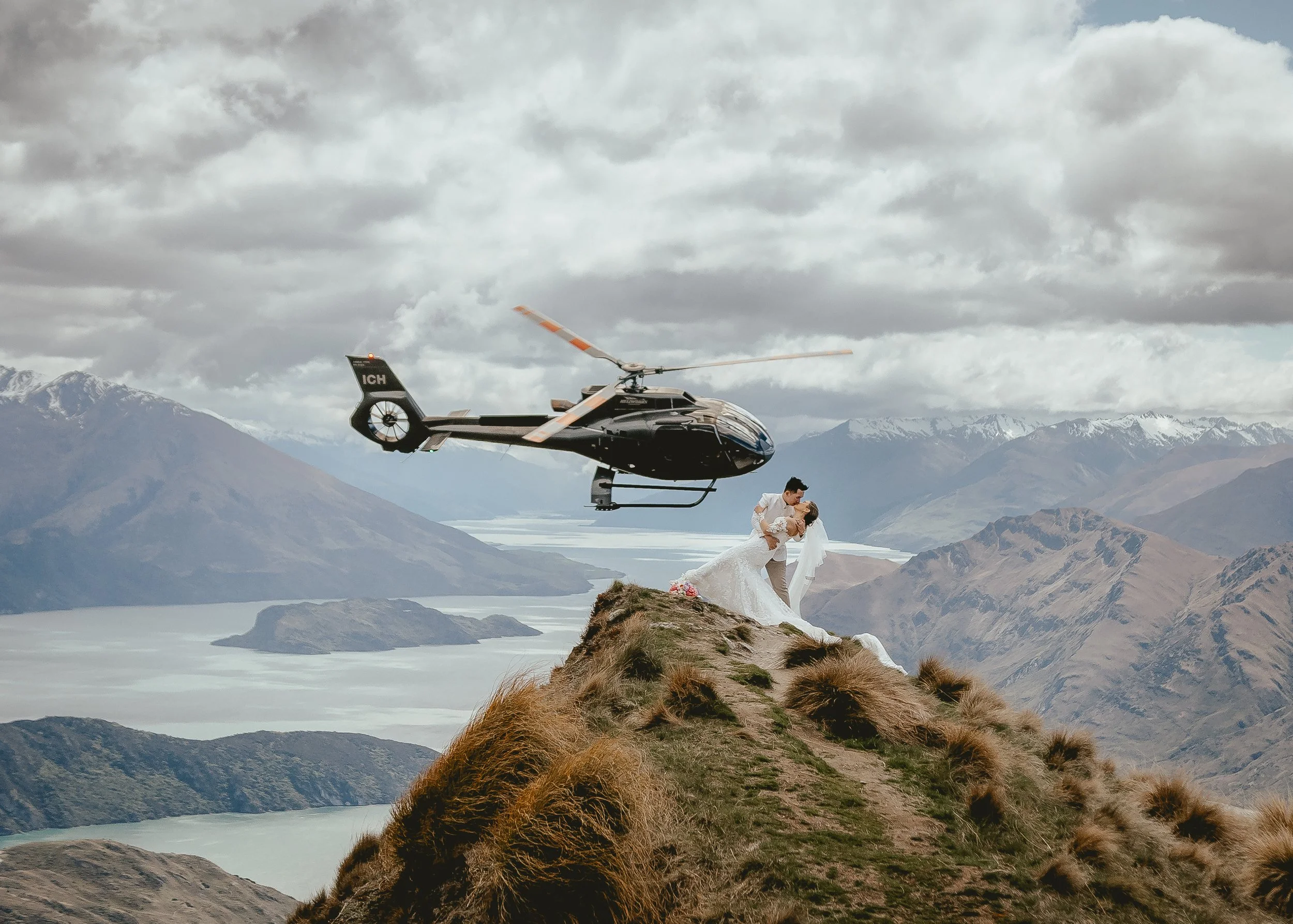 A couple in wedding attire standing on a rocky hilltop in a mountainous landscape with a helicopter flying overhead.