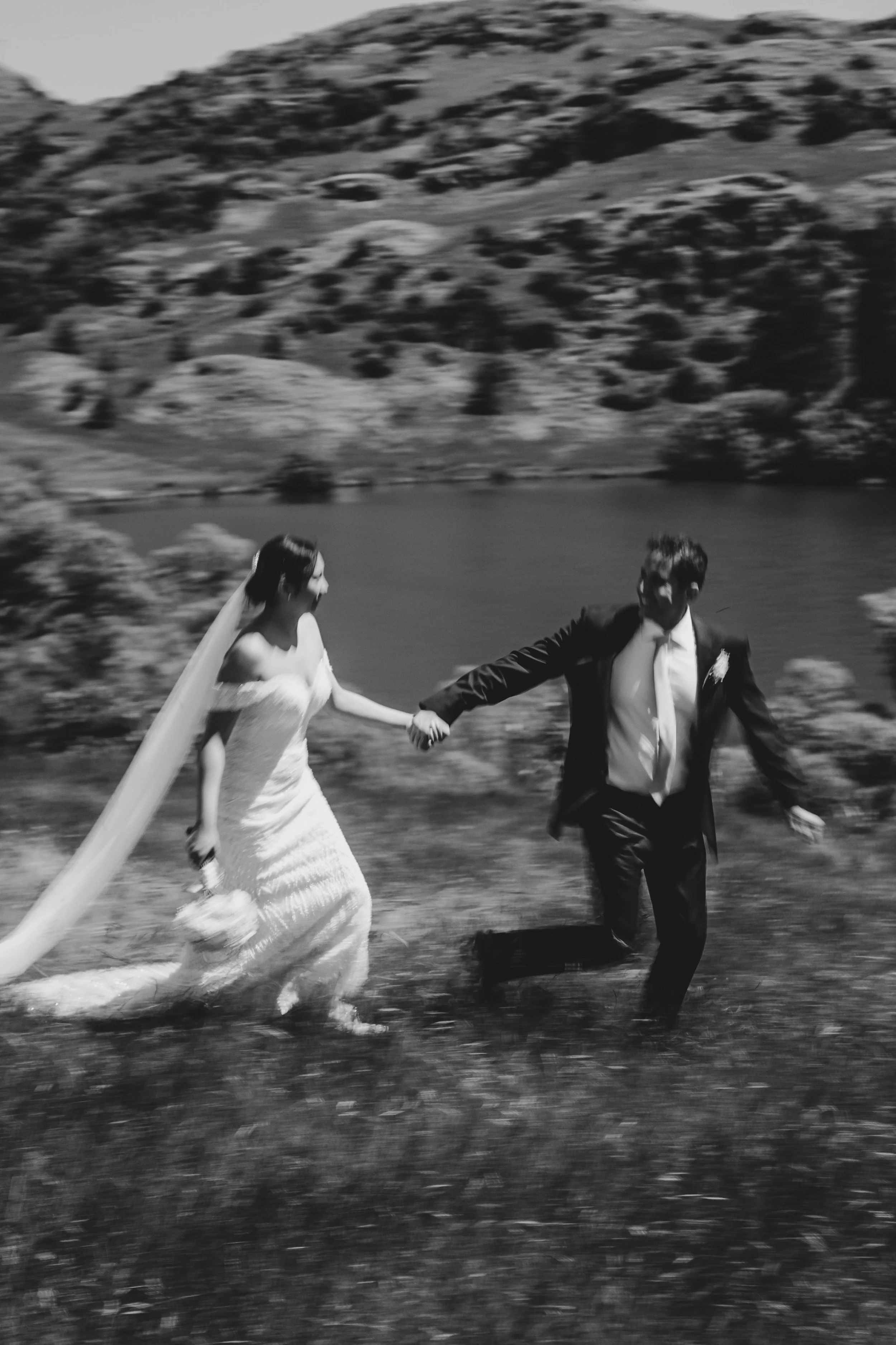 Black and white photo of a bride and groom holding hands and running outdoors with mountains and trees in the background.